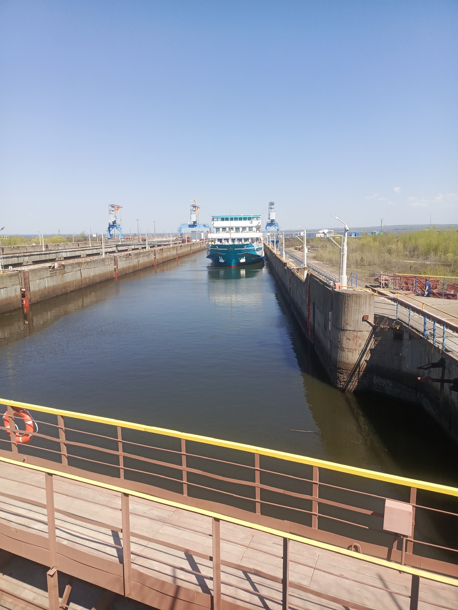 The image shows a ship in a lock on the Kama River, likely in the city of Chaikovsky, Russia, where there is a significant river port and canal infrastructure. The ship is teal and white and appears to be a passenger or transport vessel. The lock is constructed of concrete walls. The water is dark and still. Overhead, there are industrial cranes on both sides of the lock. The weather is sunny and the sky is clear. A pedestrian walkway with a yellow-railed fence runs along the near side of the lock. A life preserver is visible on the railing. Distant trees are visible on the shoreline.