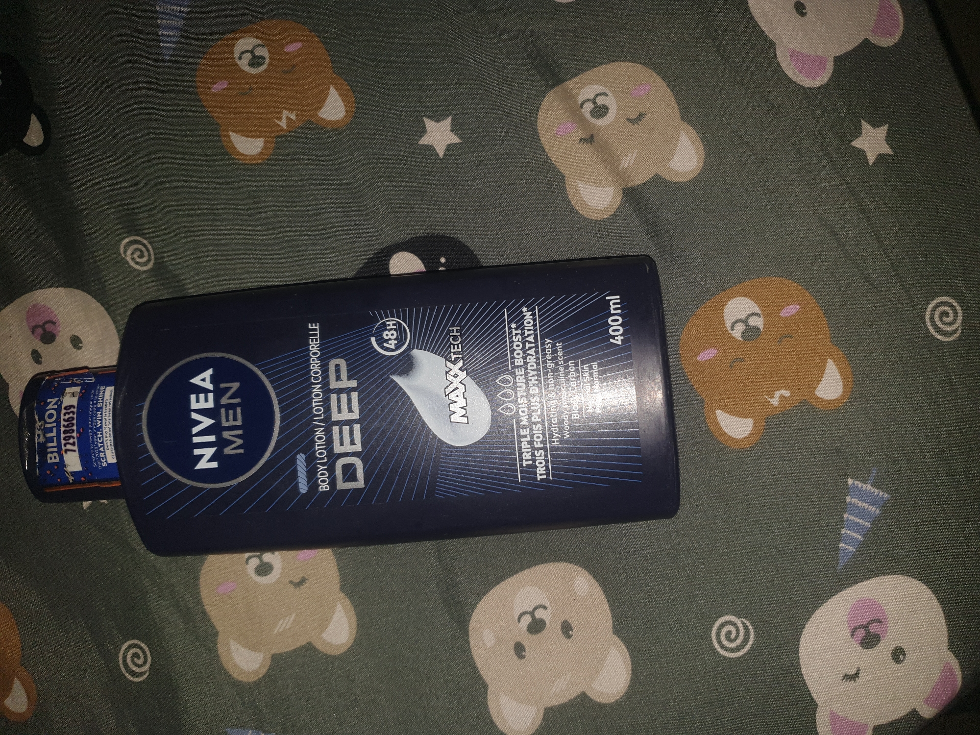 A close-up, top-down shot features a dark blue bottle of NIVEA MEN DEEP Body Lotion resting on a grey-green fabric with a pattern of cartoon bears and stars. The bottle is positioned slightly off-center to the right, with its cap facing upwards. The label is clearly visible, displaying the NIVEA MEN logo, "DEEP" in large silver letters, and smaller text detailing "BODY LOTION / LOTION CORPORELLE". Below this, text indicates "TRIPLE MOISTURE BOOST" and "48H" in a circular emblem. The product volume, "400 ml", is also present at the bottom of the label. A small, sealed area with "BILLION 72986639" and instructions for a scratch-and-win promotion is attached to the cap. The fabric beneath the bottle has a repeating pattern of stylized bears in various colors, including light brown, white with pink accents, and a brown bear. Several small white stars and swirl designs are also scattered across the fabric. The lighting suggests an indoor setting, with no discernible city or environmental context beyond the product and its immediate surroundings. The scene does not provide any information about the location in Jalingo, Nigeria.