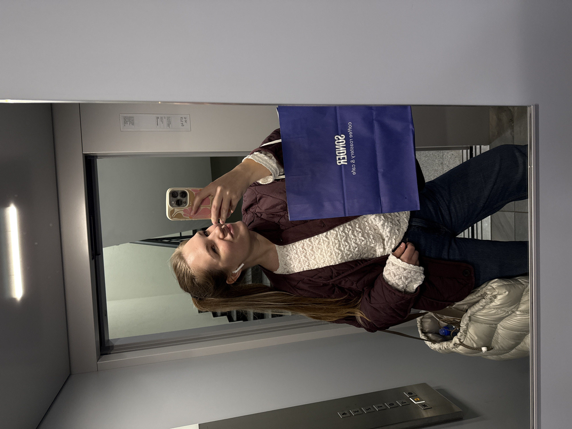 A young woman with long, light brown hair is taking a selfie in an elevator. She's wearing a white, lacy top under a burgundy jacket and dark jeans. She holds a smartphone in her left hand, and a dark blue shopping bag obscures part of her face. An earpiece is visible in her left ear. A silver puffer bag is near her. The elevator's control panel is visible, along with the elevator door. The reflection shows the elevator hallway and some architectural details. It's not possible to identify the exact location in the city of Vozdovac Urban Municipality, Serbia, based on the provided image.