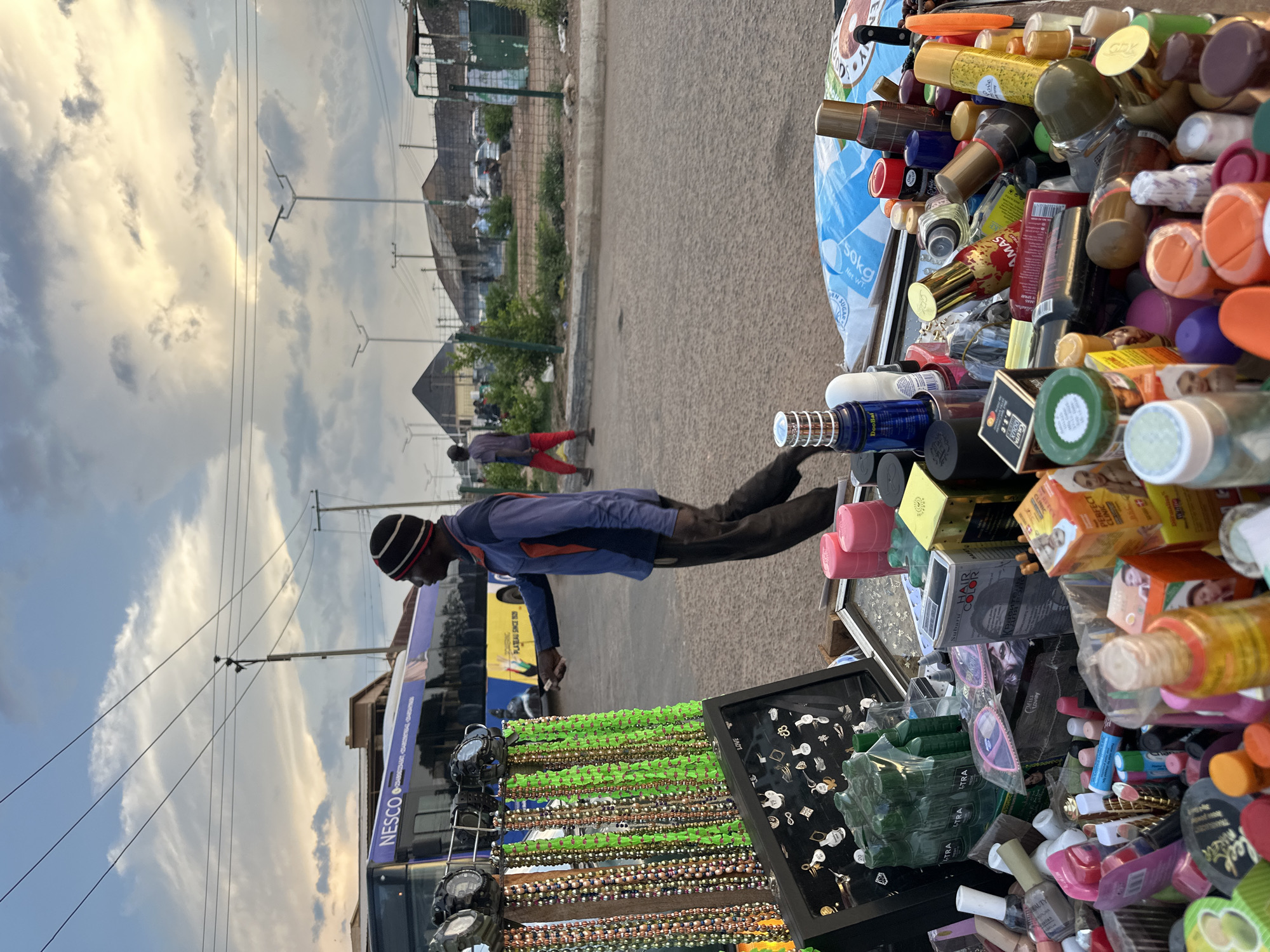 The image depicts an outdoor market stall in Bukuru, Nigeria, with a vendor and customers. The stall displays a variety of items, including necklaces, rings, perfumes, cosmetics, and hair products. A man, likely the vendor, is wearing a striped beanie and a blue and orange long-sleeved shirt, standing behind the stall and facing left, with his hand outstretched. Another person, wearing red pants and a dark shirt, walks away from the stall in the background. A NESCO bus is parked behind the stall, and other vehicles and structures are visible in the distance. The sky is cloudy, suggesting it may be late afternoon or early evening. The overall scene is of a busy market environment.