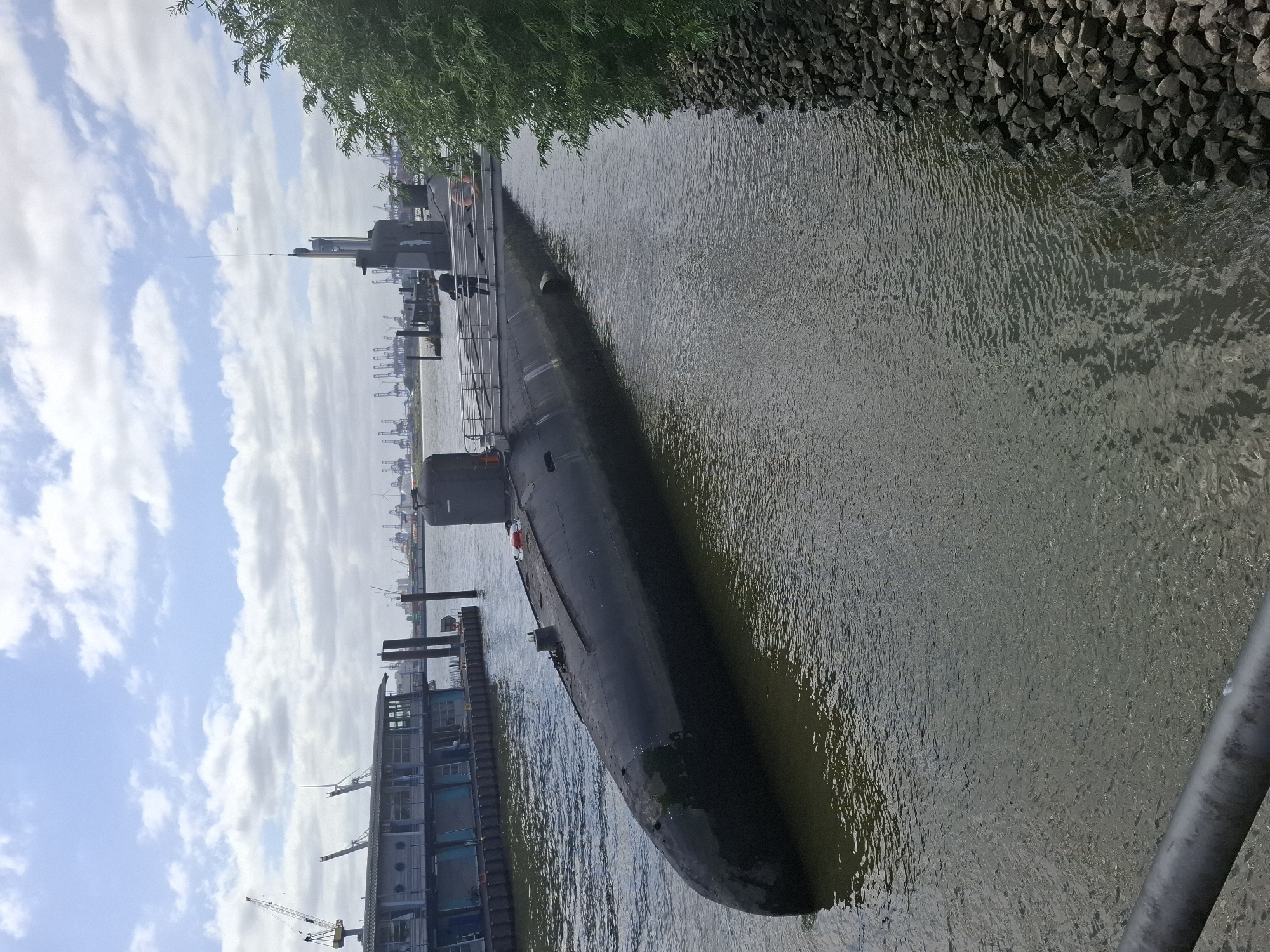 The image shows a submarine docked in a harbor in Hamburg, Germany. The submarine is dark gray and partially submerged in the water. There is a walkway leading to the submarine, and a person is standing on the walkway. In the background, there are cranes and other ships. The sky is cloudy.