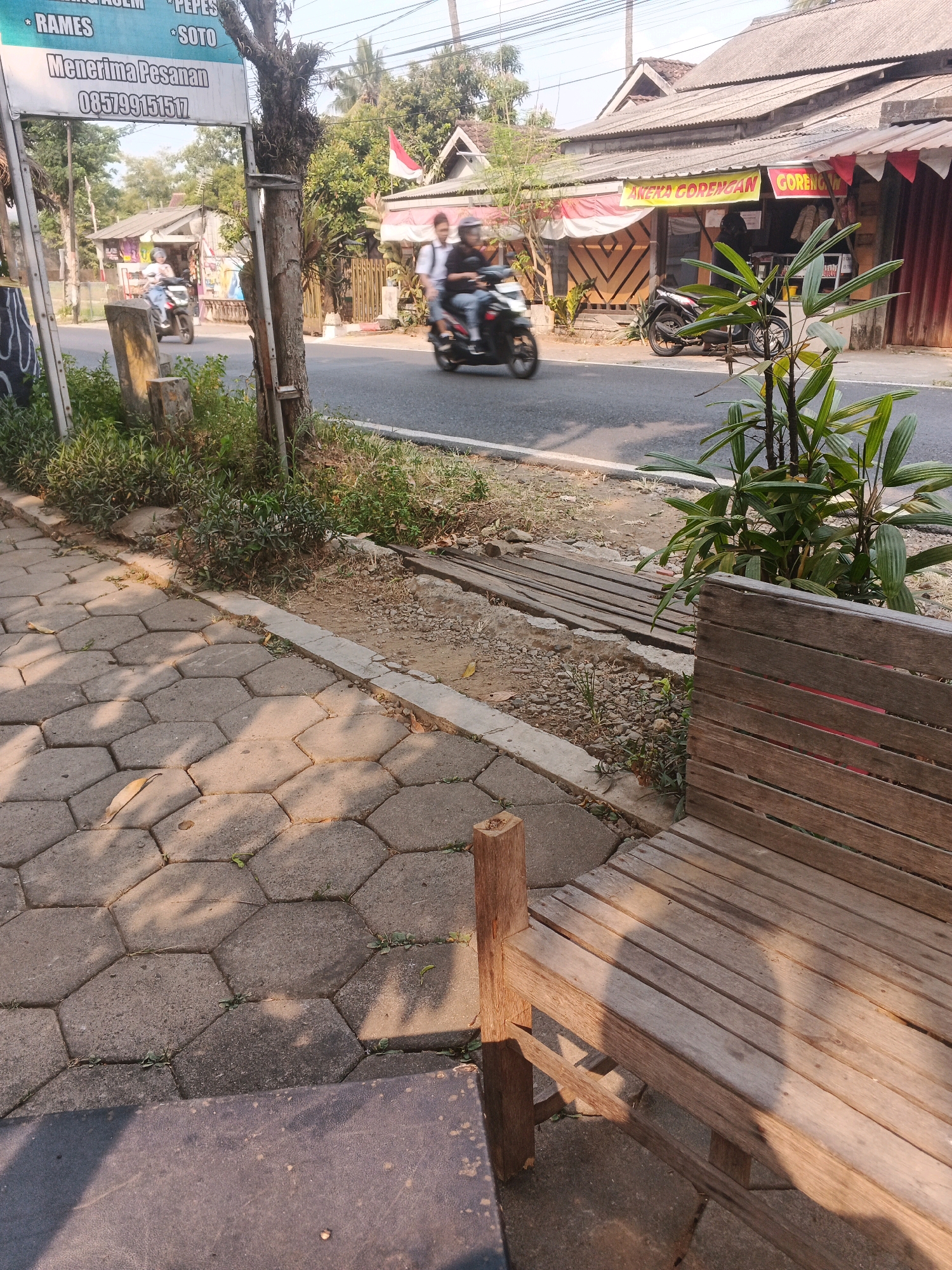The image depicts a street scene in Ungaran, Indonesia, on a sunny day. In the foreground, a wooden bench sits on a hexagonal paver sidewalk. The sidewalk is partially shaded by trees. To the right of the bench, a young palm plant grows. The street is visible in the middle ground, with a few motorcycles and riders passing by. On the left side of the street, a sign reads "RAMES * SOTO" and "Menerima Pesanan 0857991517". Further down the street, on the right, is a building with a red and white awning and signs that appear to advertise food, possibly "ANEKA GORENGAN" and "GORENG". The overall impression is of a quiet, somewhat rustic street in a town.