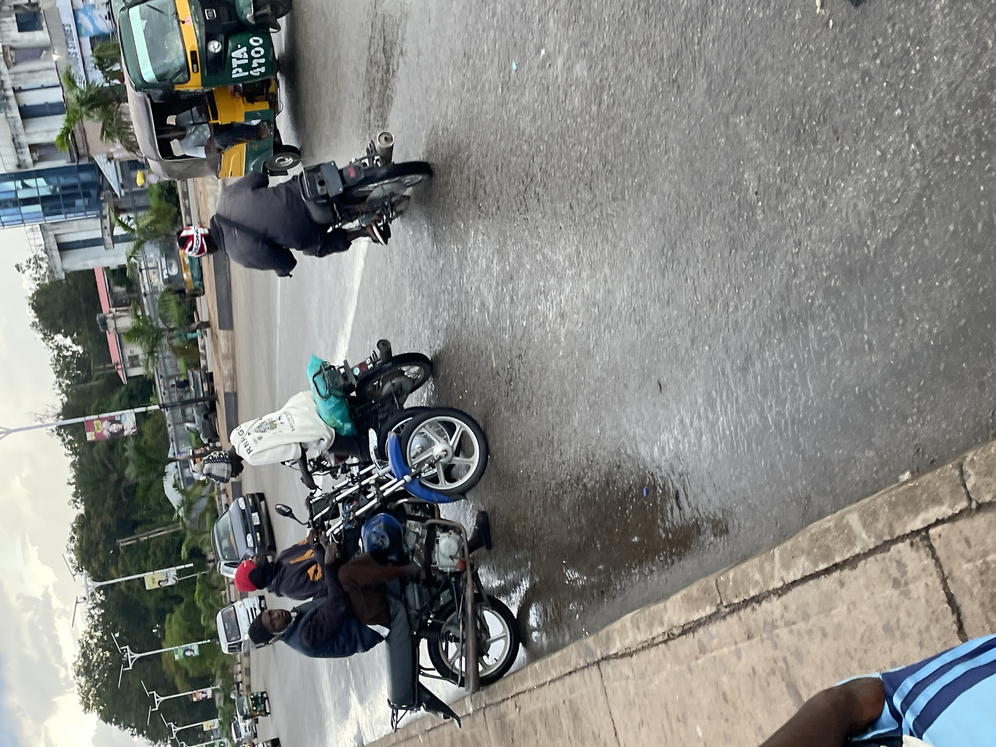 The image captures a street scene in Barakin Lerawi, Nigeria, with several motorcycles and a motorized rickshaw (tricycle) on a wet asphalt road. In the foreground, two men are seated on a blue motorcycle. The driver, on the left, wears a dark jacket and brown trousers. The passenger, on the right, has a red head covering and a yellow and black jacket. Behind them, another motorcycle with a rider wearing a white garment is visible. To the right, a green and yellow tricycle is in motion, with a person seated inside. The background features buildings, trees, and streetlights, some with solar panels. The ground is wet, indicating recent rain. The overall atmosphere suggests an active urban environment.