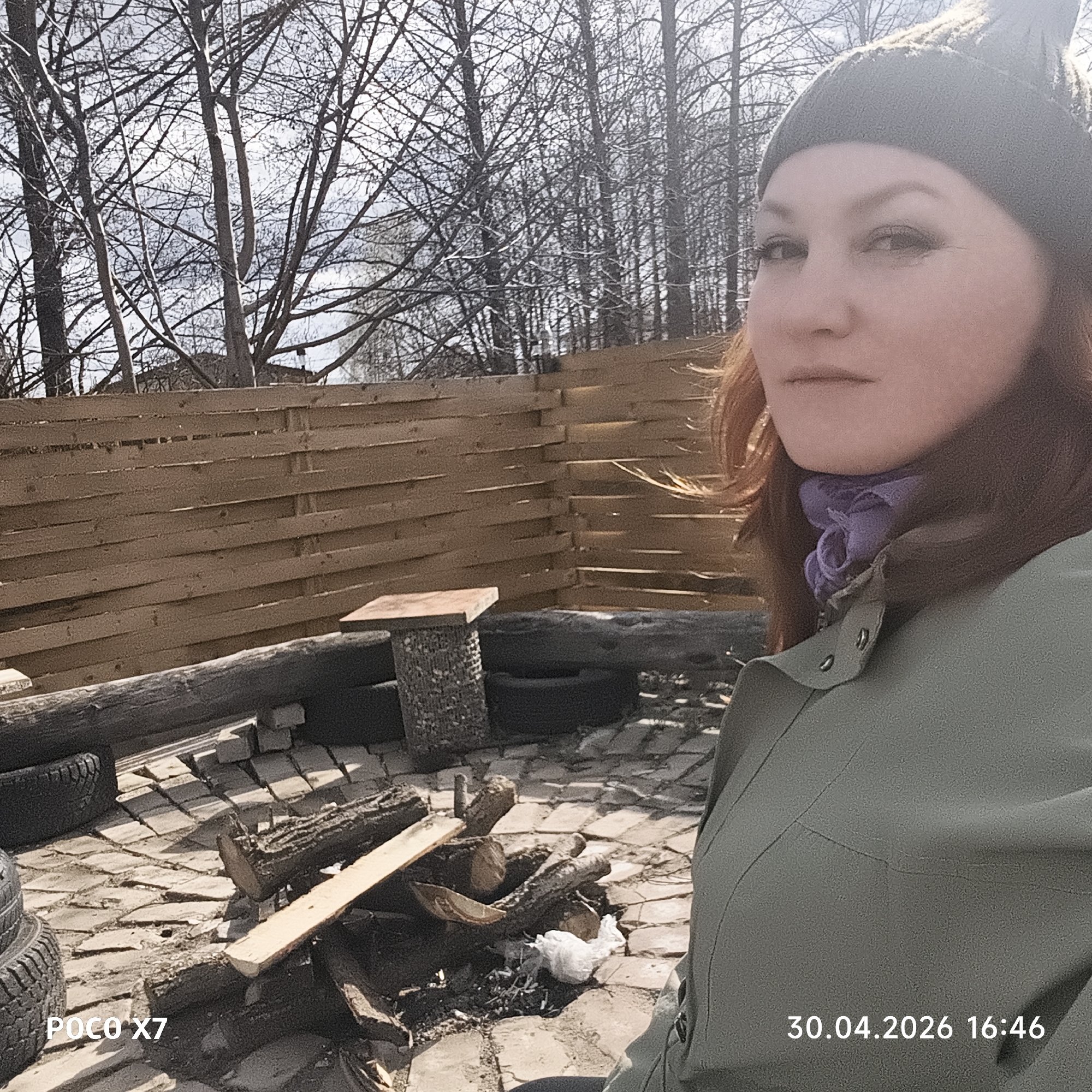 The image features a woman posing for a photograph in an outdoor setting in Chaikovsky, Russia. The woman, with reddish-brown hair, is positioned in the right foreground, wearing a dark green beanie, a light purple scarf, and an olive green jacket. Her expression is neutral as she looks towards the right.
The scene depicts a rustic outdoor area with a brick or paver ground. Visible objects include several cut logs and wooden planks, possibly for a campfire, and repurposed tires arranged on the ground. A small, makeshift table with a wooden top and a stone-like base stands in the midground, with a white plastic bag lying near the logs. A wooden slatted fence extends across the midground, separating the immediate area from the background. Behind the fence, numerous bare trees with intricate branches are silhouetted against a bright, possibly cloudy, sky, suggesting late winter or early spring conditions.
The overall lighting indicates daytime. Visible text includes "POCO X7" in the bottom left corner and "30.04.2026 16:46" in the bottom right corner, specifying the camera model and the date and time of capture as late afternoon.