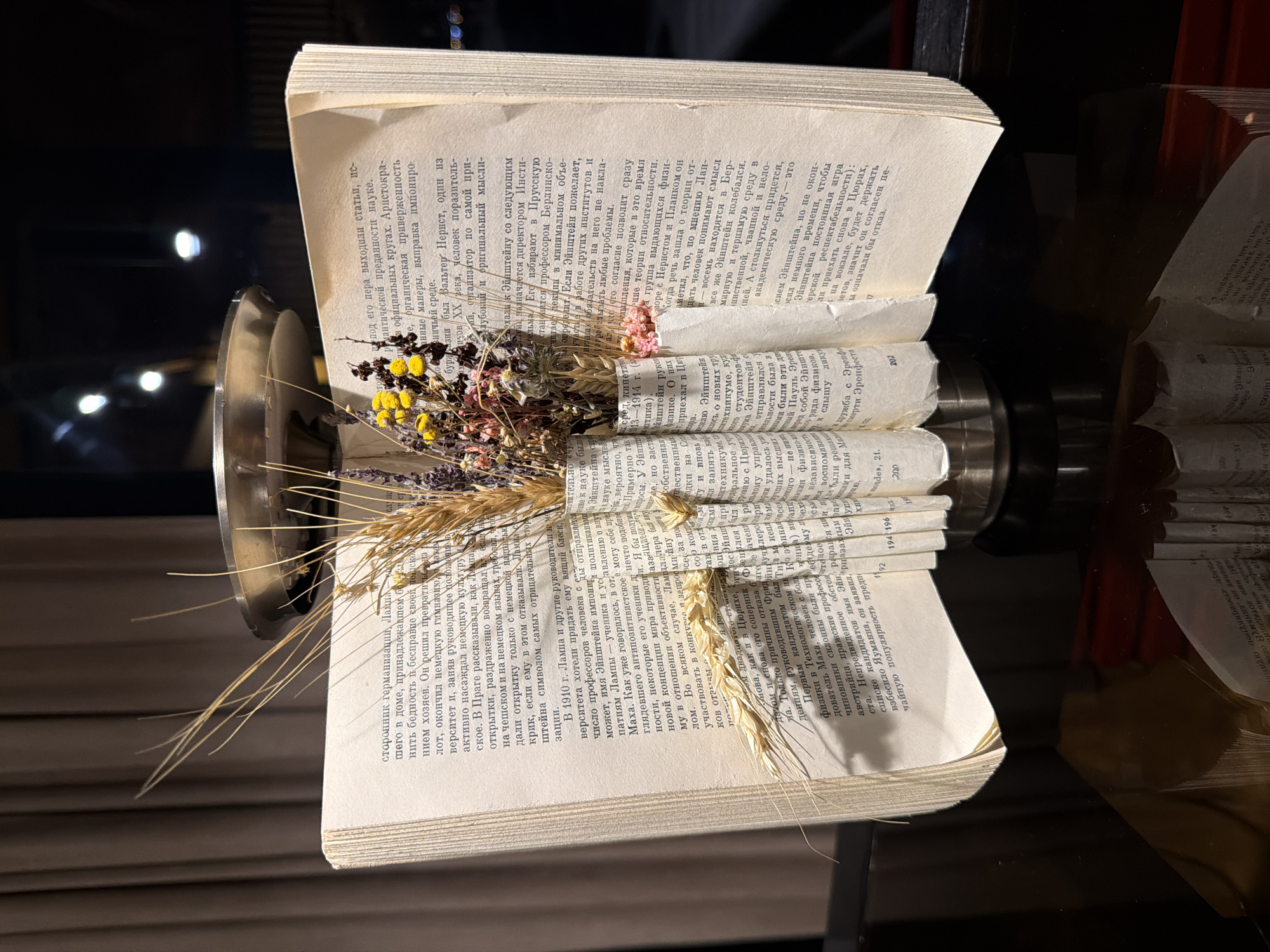 The image shows an open book with pages that have been rolled up around a metal cylindrical object, and a small dried flower arrangement placed on top of the rolled pages. The book has text written in a Cyrillic script. The metal cylinder seems to be supporting the rolled pages and floral arrangement. The background appears to be a dark, possibly dimly lit environment, with reflective surfaces suggesting a table or counter.