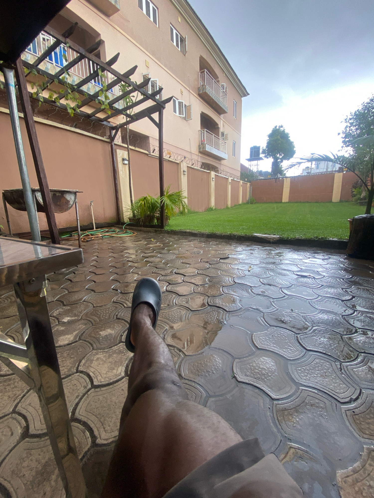 In Barden Yana, Nigeria, a man's leg, clad in a black slipper, rests on a wet, patterned paved patio. The patio glistens with puddles, reflecting the overcast sky and a two-story building with cream-colored walls and balconies. To the left, a metal table with a glass top is partially visible. Beyond the patio, a lush green lawn stretches towards a brown fence. A wooden pergola with climbing vines and a barbecue grill are also present. In the distance, trees and a water tank can be seen, with the general atmosphere suggesting a rainy day.