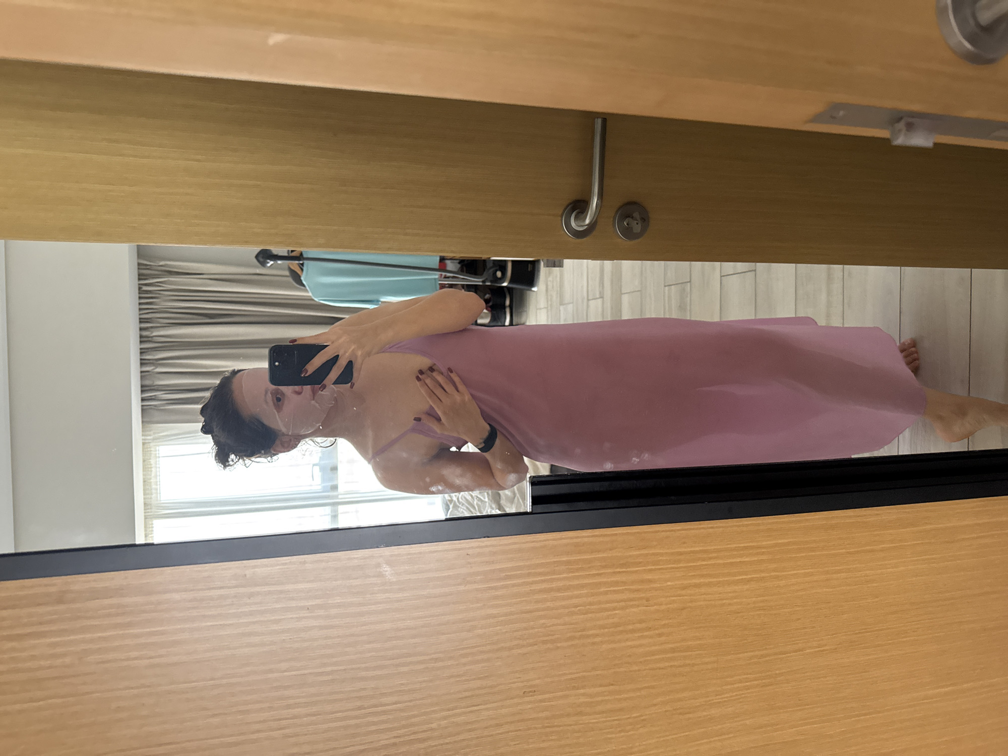 The image shows a woman in a pink garment taking a selfie in a mirror. She is holding a smartphone and has a beauty mask on her face. The reflection shows a room interior with curtains, a suitcase, and tiled flooring. The location is suggested to be in Dubai, United Arab Emirates. The mirror is framed by a wooden structure, possibly a door or wardrobe.