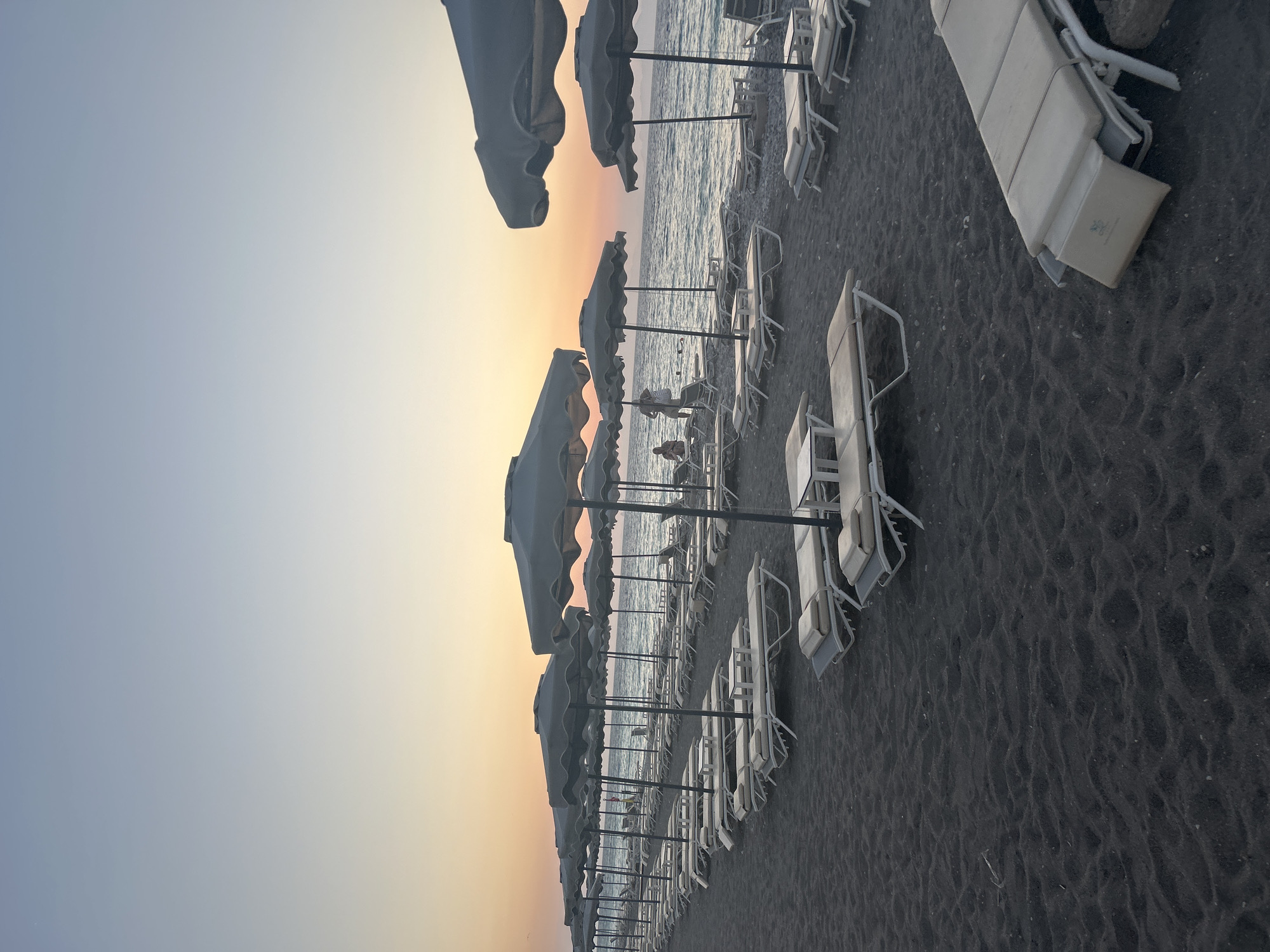 This is a vertically oriented, eye-level photograph of a black sand beach in Rhodes, Greece, during either sunrise or sunset. The scene is dominated by rows of empty white sun loungers and closed umbrellas, arranged neatly on the dark, textured sand. The calm sea stretches out in the background, meeting a pale, gradient sky that shifts from a soft yellow and orange near the horizon to a light blue overhead.
Several figures, appearing as small silhouettes, can be seen near the water's edge, suggesting a few people are enjoying the beach despite the apparent early or late hour. The overall mood of the image is tranquil and serene, with the quiet beach and the soft glow of the sky creating a peaceful atmosphere. The sun loungers are made of a light-colored material, possibly canvas or a similar fabric, and are set on white metal frames. The umbrellas are closed, their fabric a muted gray or off-white. The black sand is uneven, showing natural ripples and indentations. A single visible logo on one of the sun loungers appears to be a circular emblem with some text, but it is too indistinct to be read.
