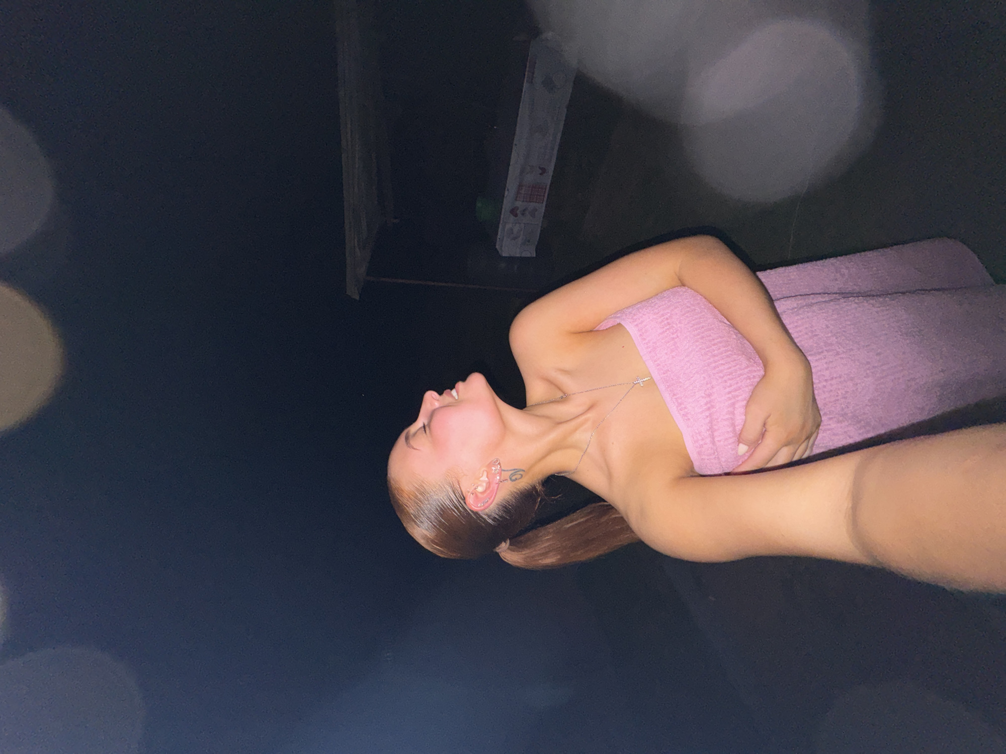 A vertical indoor shot shows a woman posing in Chastyye, Russia. The woman has brown hair pulled back in a low ponytail and is wearing a pink wrap. She is on a dark, smooth surface and is facing up and to the left. There is a white rectangular object with red hearts on it in the top left corner. There are white, out-of-focus spheres in the image. The woman has earrings and a tattoo on her neck.