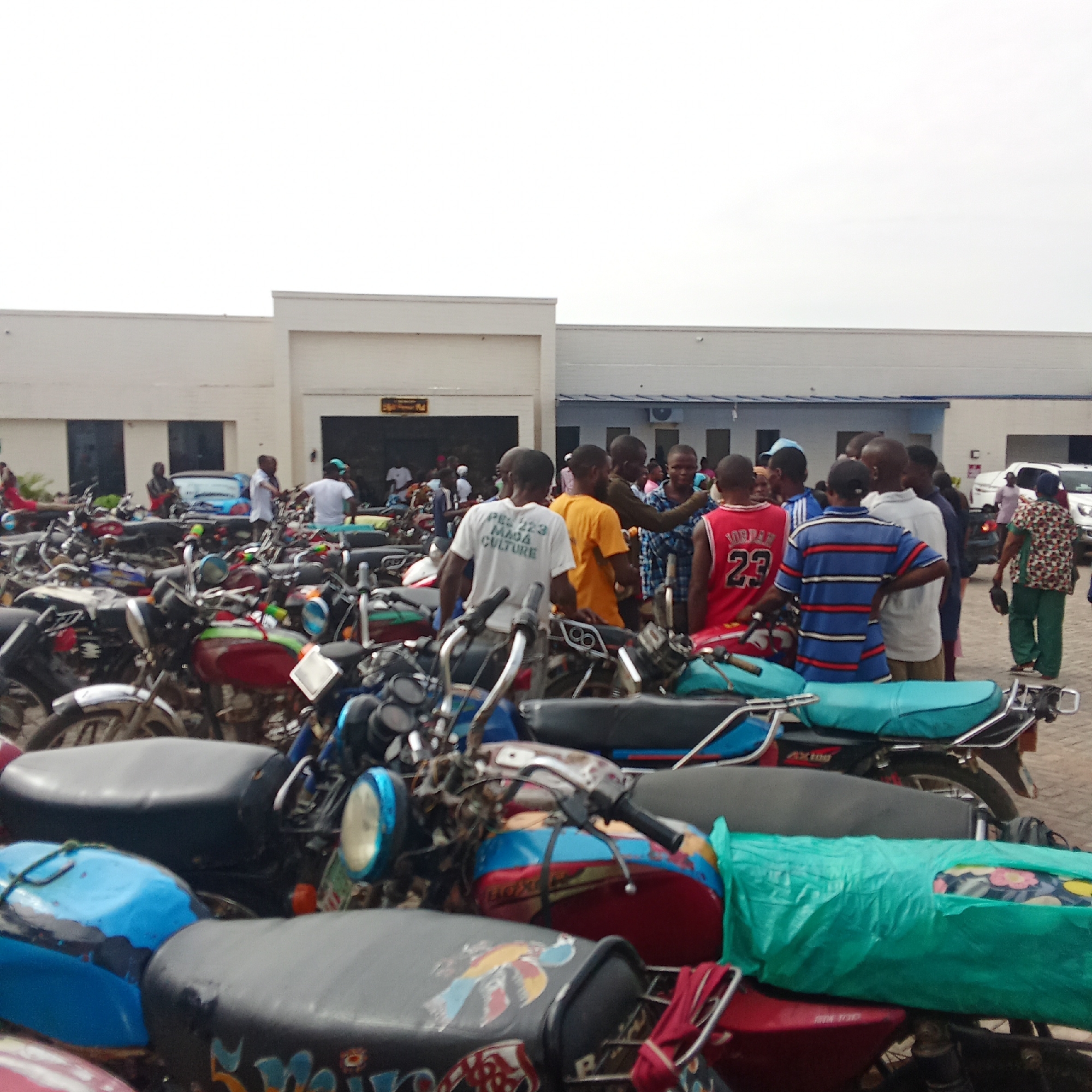 The image depicts a crowded scene outside a building, likely a government or public facility in Akwanga, Nigeria. The foreground is dominated by numerous motorcycles, many appearing in a state of disrepair, parked haphazardly. A group of people, primarily men and young boys, are gathered near the building's entrance. They are dressed in casual clothing, including t-shirts, jerseys (one with "JORDAN 23" visible), and patterned shirts. A woman in green pants and a patterned top is visible on the right side of the image. In the background, a white vehicle, possibly a car, is parked near the building. The building itself is a low, white structure with a visible sign above the entrance. The overall impression is one of congestion and activity in a public space.