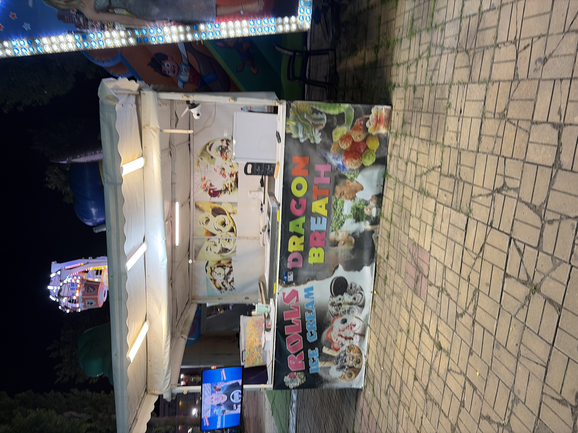 An outdoor shot captures a food stall at night, displaying "ROLLS ICE CREAM" and "DRAGON BREATH" signage with colorful graphics. The stall is illuminated by overhead lights and a vertical string of blue lights on the right. Inside the stall, various ice cream flavors are visible on display. To the left of the stall, a television screen shows a man wearing a black t-shirt. In the background, a Ferris wheel and other amusement park rides are faintly visible. The ground is paved with tan bricks.