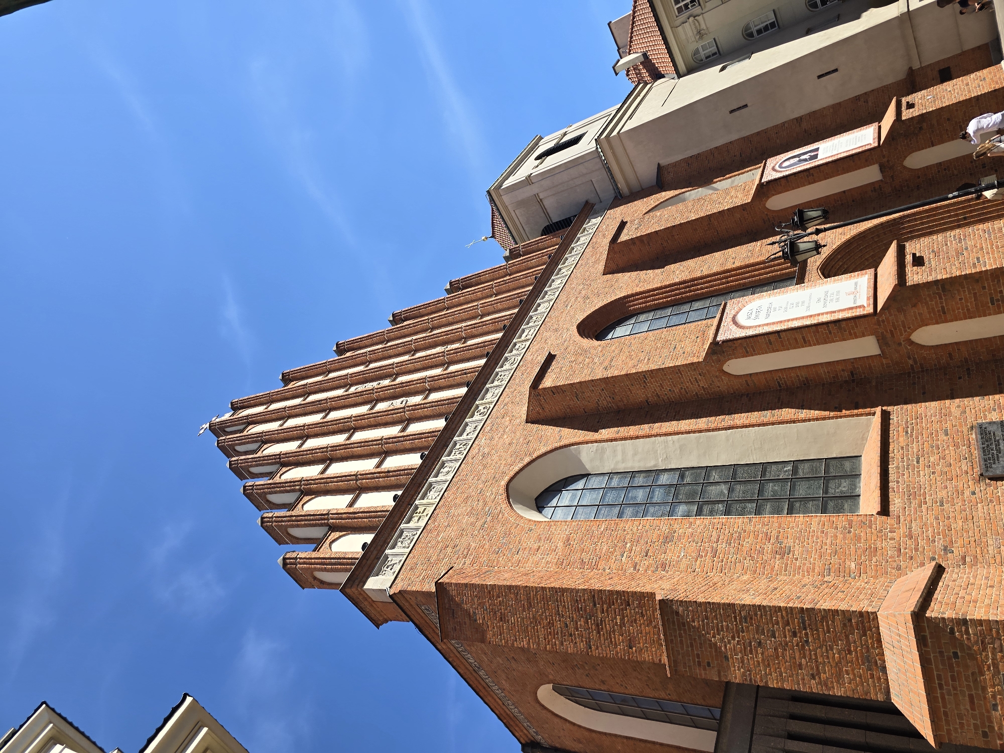 The image shows a low-angle shot of a large brick church in Warsaw, Poland. The church has a prominent steep roof with decorative elements and arched windows. A cream-colored bell tower is visible to the right of the main structure. The sky is a clear blue with a few wispy clouds. Several white banners with text and images are attached to the church facade. A street lamp stands in front of the entrance. Two people are walking on the pavement in front of the church, one man wearing a white shirt and shorts, and a woman in a light-colored dress. The architectural style suggests it is a historic Gothic Revival or Neo-Gothic building, commonly found in European cities. The scene is bathed in bright sunlight, indicating a daytime shot.