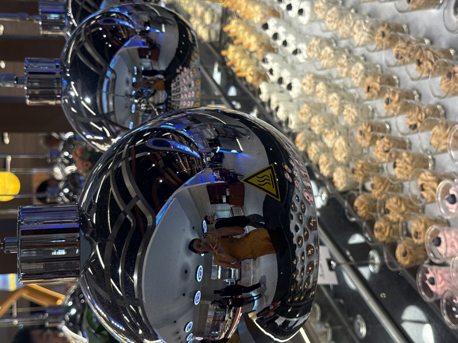 Two silver, spherical objects with cylindrical attachments reflect the surrounding environment. Reflections include people, a restaurant setting, and a warning symbol. A grid of small glass cylinders containing light brown and white objects is visible in the background. The cylinders appear to be arranged in rows and columns, possibly as a display or decoration. The objects have a consistent shape with circles on the front.