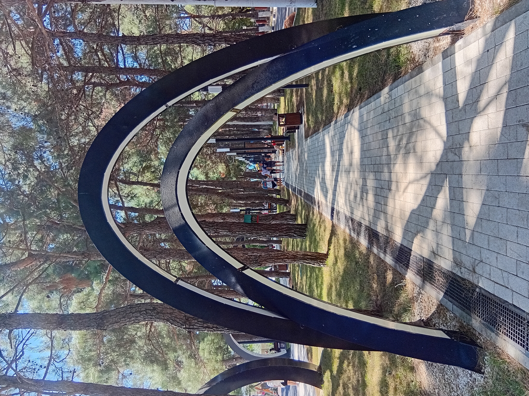 The image shows a park pathway lined with trees. In the foreground, two large, dark, arched structures are visible, framing the pathway. The pathway itself is paved with light-colored rectangular stones and is illuminated by sunlight casting shadows. Several people are walking on the pathway further ahead. The environment is lush with green grass and tall trees. There are lampposts along the side of the path. In the background, there are more trees and some buildings or structures that are not clearly discernible. The scene appears to be a public park or promenade.