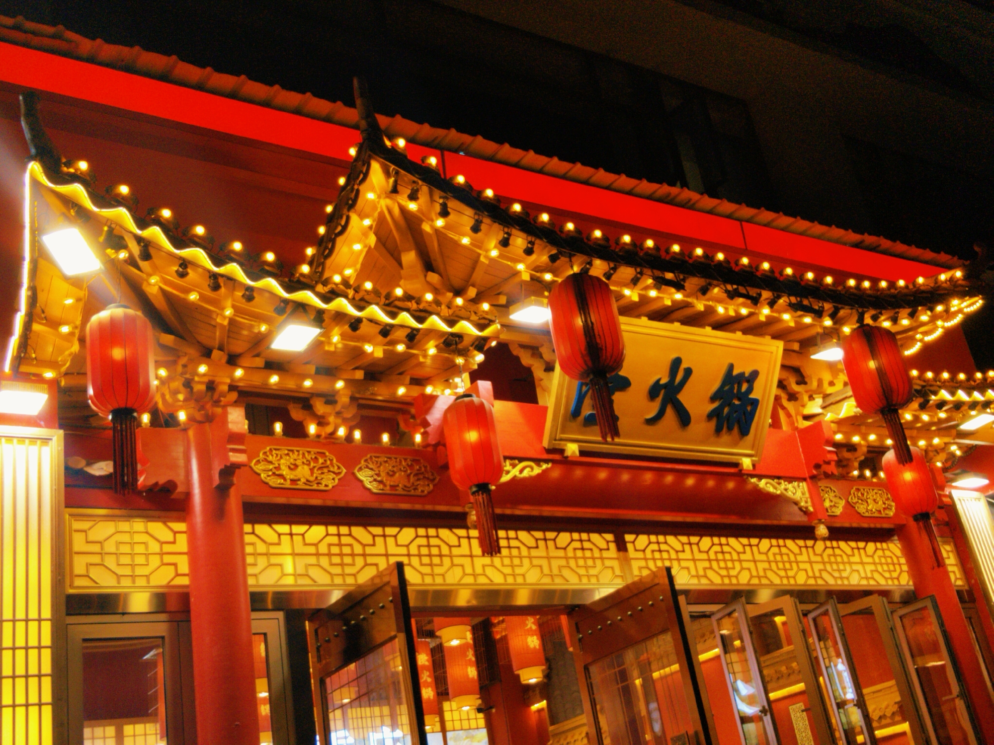 Captured at night in Yuzhong District, China, the image showcases an illuminated Chinese restaurant. The architecture features a red roof, ornate gold trim, and traditional Chinese lanterns. Strings of lights accent the eaves. A signboard with Chinese characters is prominently displayed. Open doors reveal the interior, reflecting the warm light of the establishment. The composition is from a low angle, emphasizing the height and grandeur of the building.