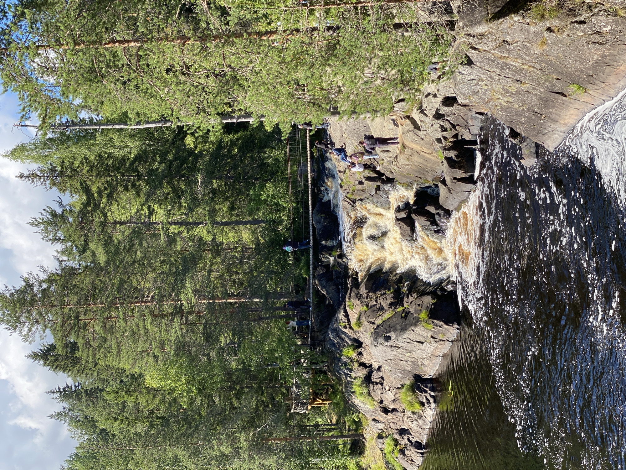 The image depicts a scenic waterfall in Ruskeala, Russia, a town renowned for its marble canyons. A tiered waterfall cascades down dark, rocky cliffs into a dark, rippling body of water in the foreground. Several people are visible in the scene, some standing on the rocks near the waterfall, others on a suspension bridge that spans the river upstream. The bridge is made of wood and rope, with wooden railings. A wooden viewing platform is also visible to the left of the bridge. The surrounding environment is a dense forest of tall pine trees and other deciduous trees under a partly cloudy sky. The water is a murky brown, likely due to sediment from the waterfall, and white foam is visible on its surface. The rocks are rugged and uneven, covered in patches of green moss and grass. The overall impression is one of a natural, rugged landscape enhanced by man-made structures for visitor access.