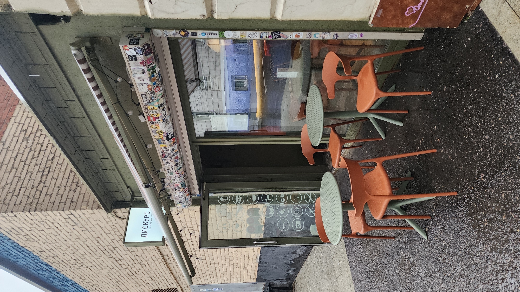 Outside a building in Moscow, Russia, a sidewalk cafe features round, green tables and orange, modern-style chairs. The building has a green awning covered in stickers. A sign in Russian hangs beneath the awning. The window of the building has stickers. The ground is wet.