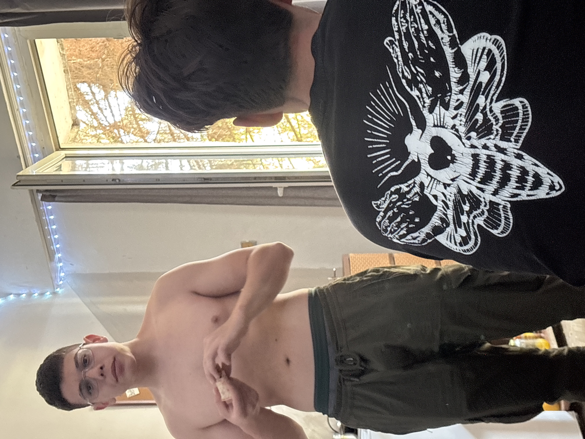 An indoor scene, rotated 90 degrees clockwise, features two individuals. In the foreground, a young man, shirtless and wearing glasses, looks directly towards the camera. He has dark, short hair and is wearing olive green cargo pants with dark underwear visible at the waistband. His hands are clasped in front of him, holding and seemingly tearing a small, light-colored object, possibly a piece of bread or pastry. His expression is neutral.
Behind and to the right of this man, the back of another person's head with dark hair and their right shoulder are visible. This individual is wearing a black long-sleeved shirt featuring a large, intricate white graphic on the back. The graphic depicts a moth-like creature with spread wings, and what appears to be praying hands forming its body or head, with rays radiating from behind it.
To the left, a bright window or skylight is open, revealing a view of dense, bare tree branches, suggesting a cold season like winter or early spring. String lights with small white bulbs are strung along the wall or window frame near the shirtless man. A subtle hint of wicker furniture or a basket is visible in the background on the right. The overall setting appears to be a casual indoor space in Troyan, Bulgaria, with natural light indicating daytime. No discernible text is visible in the image.