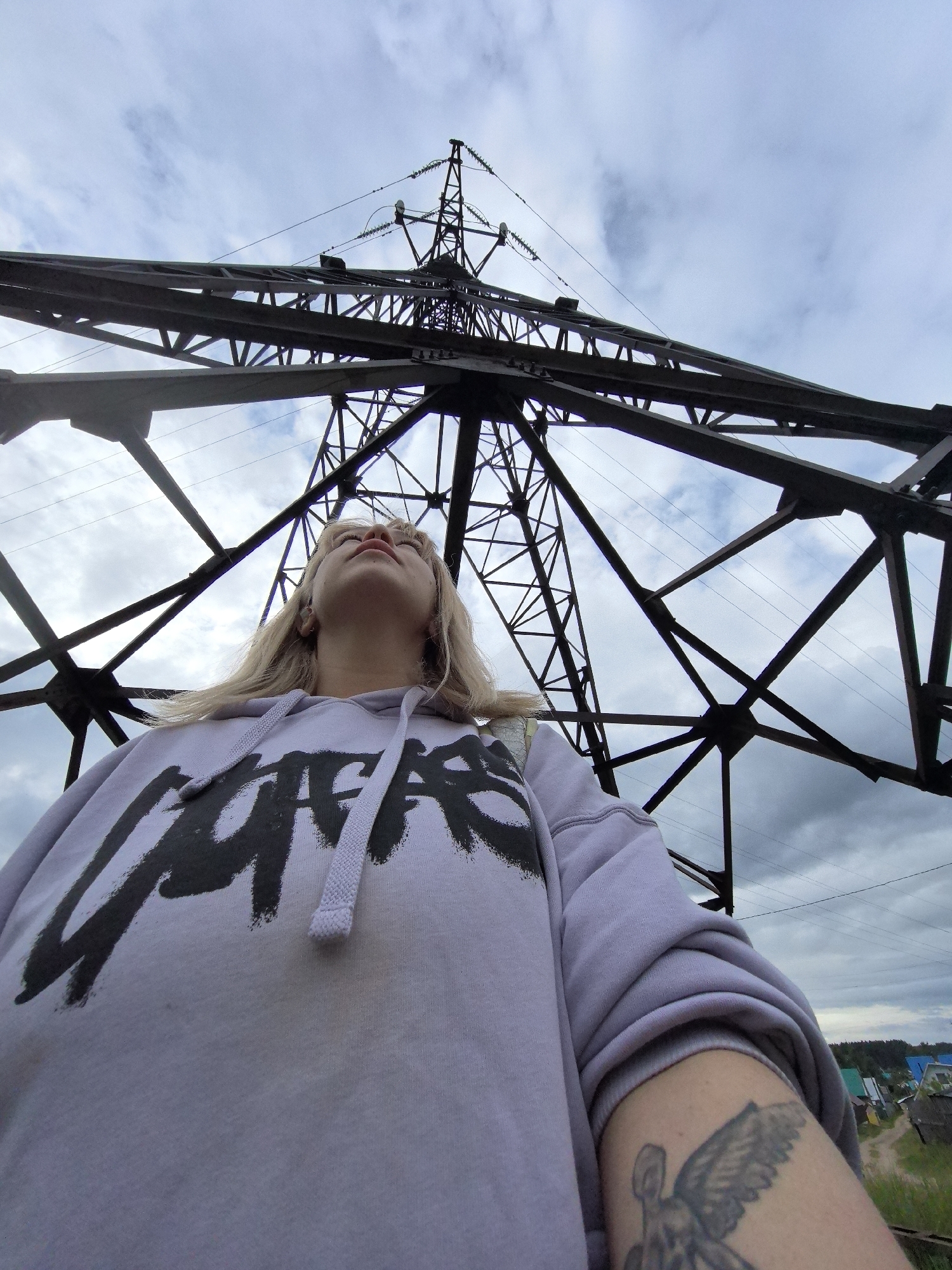 A young person, likely female with blonde hair, is captured from a low-angle perspective, looking upwards directly beneath a massive electrical transmission tower. The intricate black metal framework of the tower dominates the upper and central areas of the image, creating a strong diagonal and vertical composition against an overcast sky filled with scattered clouds.
The subject's head is tilted back, gazing towards the top of the towering structure. She wears a light purple hooded sweatshirt featuring a large, stylized dark graphic print that appears to read "STUSSY." On her right forearm, a visible tattoo depicts an angel or winged figure. Her expression is neutral and contemplative.
In the background, beyond the tower's structure and the subject, glimpses of a rural or semi-rural setting can be seen. Small houses with colorful roofs are nestled among green foliage, suggesting a residential area in a location like Noviy, Russia. A dirt path is also visible. The diffused light from the cloudy sky indicates a daytime setting. The photograph emphasizes the scale of human interaction with large-scale industrial infrastructure.