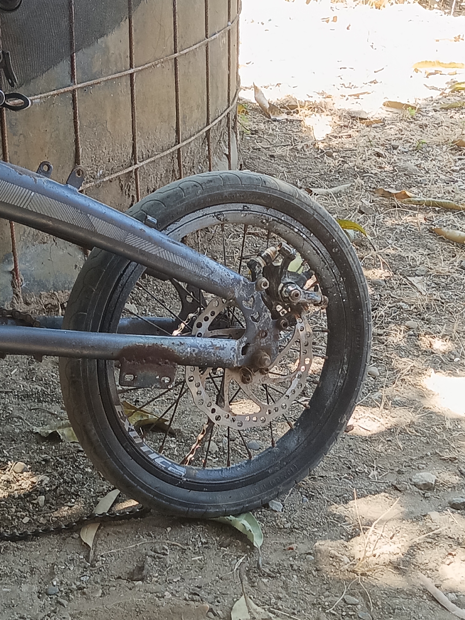 The image shows the rear wheel of a bicycle with a disc brake. The bicycle is on a dirt ground, likely outdoors, with dry leaves scattered around. In the background, a cylindrical object, possibly a water tank, is partially visible, covered with a mesh material. The overall scene suggests a dry, dusty environment, typical of a rural or outdoor setting. The location is specified as Pitue, Indonesia. There are no people or actions depicted in the image.