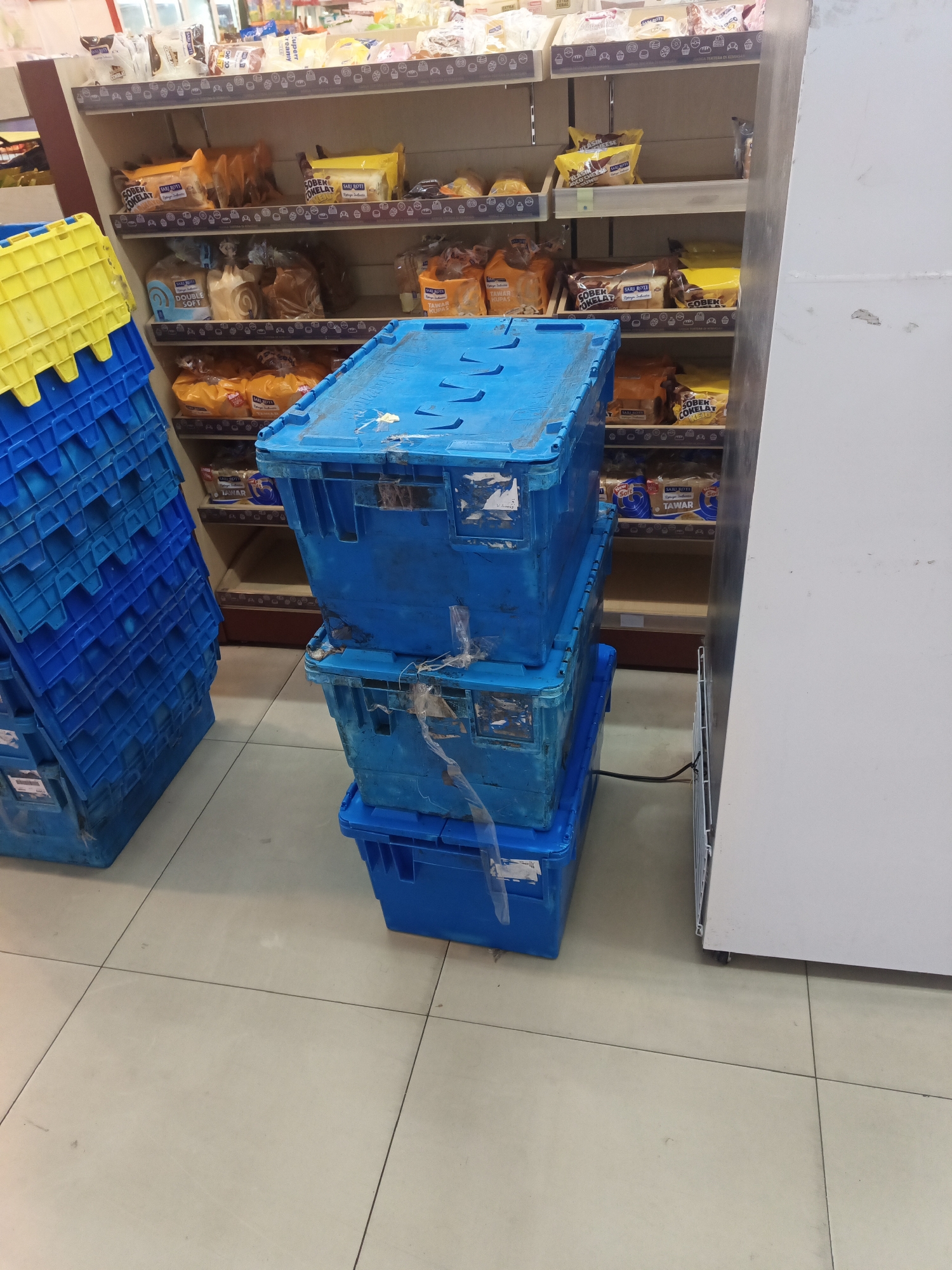 The image shows a retail environment, likely a convenience store or supermarket in North Jakarta, Indonesia, as indicated by the product packaging and general store layout. In the foreground, to the left, is a tall stack of blue plastic crates. To the right of these crates, a tower of three blue plastic containers is stacked on the floor. Behind the containers, shelves are visible, stocked with various food items, predominantly bread products in branded packaging. These include items labeled "Sari Roti" with descriptions like "Cokelat" (chocolate) and "Tawar" (plain). The overall scene suggests a stockroom or a less organized retail area. The floor is tiled, and a white refrigeration unit is partially visible on the far right. No people are present in the image.