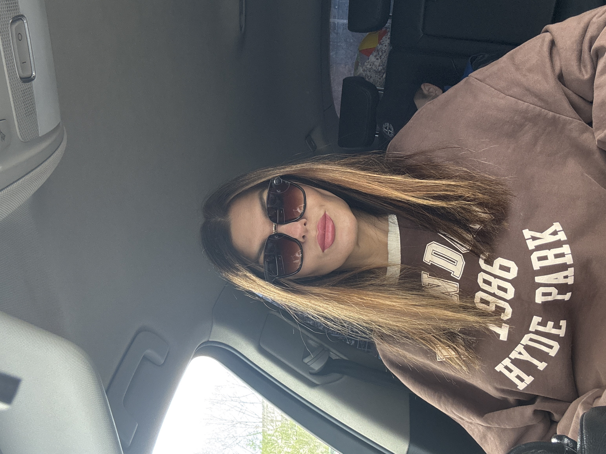 A woman is seated in a vehicle. She is wearing a brown sweatshirt with the words "HYDE PARK 1986" printed on it. She is wearing sunglasses. The interior of the vehicle is visible, including the ceiling, door, and other seats. The location is in a city of Chaikovsky, Russia. The background shows trees and buildings, suggesting the vehicle is outdoors.