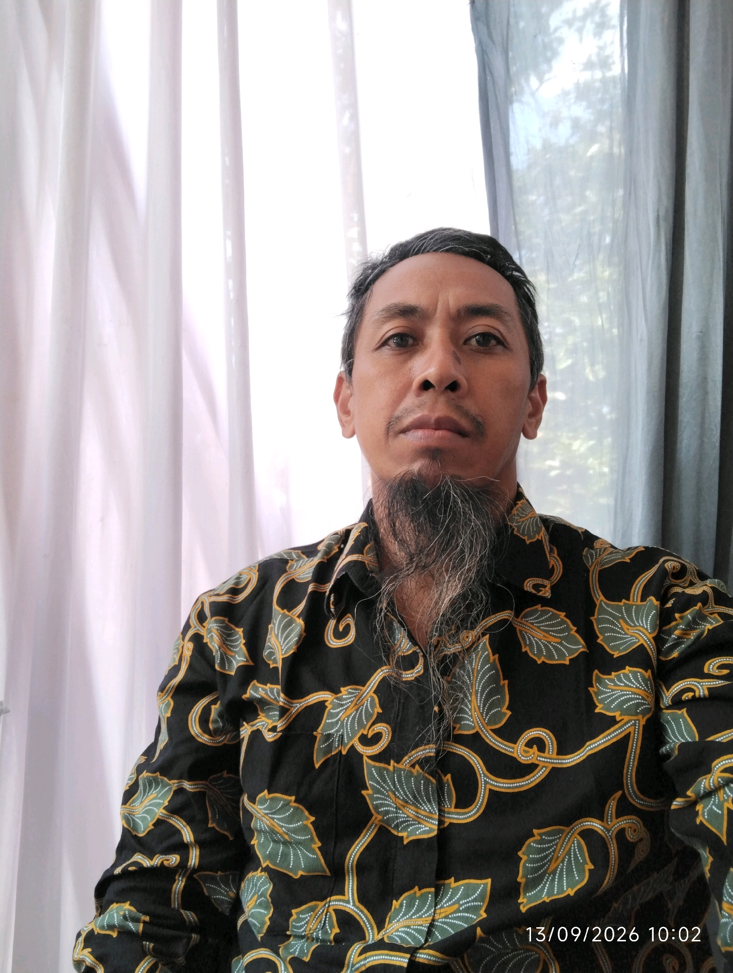 An adult male with a greying beard and mustache is pictured from the chest up, looking directly at the camera. He is wearing a black collared shirt with a gold and green leaf-like pattern. The man's left side is partially obscured by a white curtain, while a grey curtain hangs on his right side. The background suggests an indoor setting with natural light filtering through the curtains. A timestamp in the lower right corner of the image reads "13/09/2026 10:02". The location is identified as Kuwukan, Indonesia.