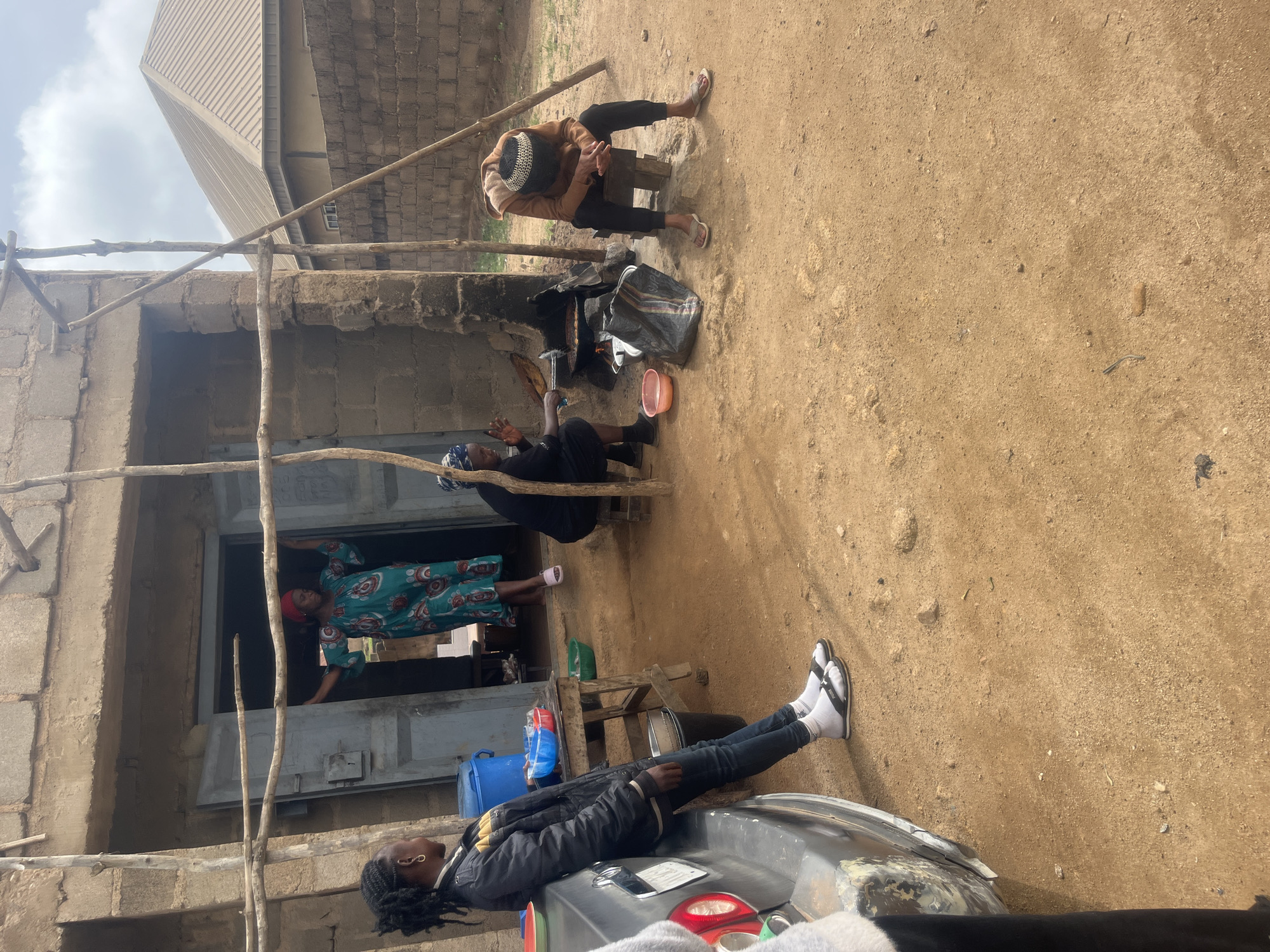 The image depicts an outdoor scene in a likely rural area. A partially constructed building with scaffolding is visible. A woman in a colorful, long-sleeved dress stands at the doorway, possibly looking out. In the foreground, an individual is lying on the hood of a car, wearing dark pants and a dark jacket. Two more people are positioned in the scene. One of them is sitting on a small stool, wearing a brown jacket and a white and black hat. The other person is seated on the ground. The ground is dirt. Without specific visual markers, confirming that this scene is from the city of Jos, Nigeria, isn't possible.