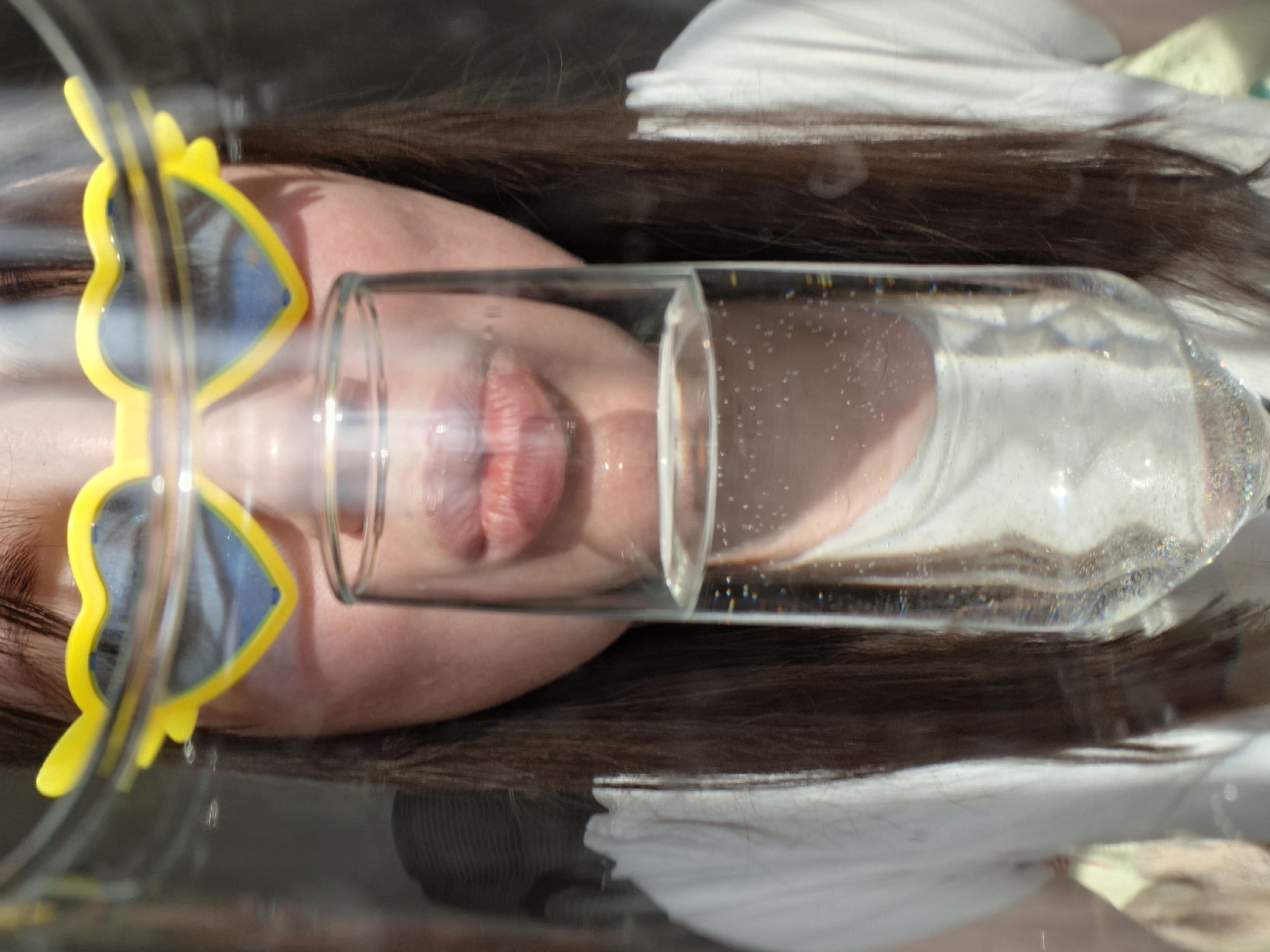 A person is lying supine with a clear glass of water resting horizontally across their face, near their lips. The person is wearing yellow sunglasses with blue lenses. The person has brown hair and is wearing a white shirt. The scene is lit by natural light. The location is Unknown, Unknown.
