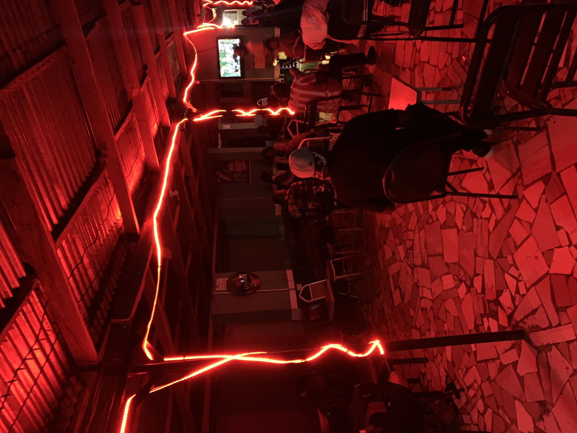 The image shows an indoor scene in what appears to be a bar or lounge. People are seated on chairs, facing a television screen mounted on the wall. Red light strips illuminate the space, accentuating the corrugated iron roof and the tiled floor. The location is presumably in or near New Stadium, Nigeria.