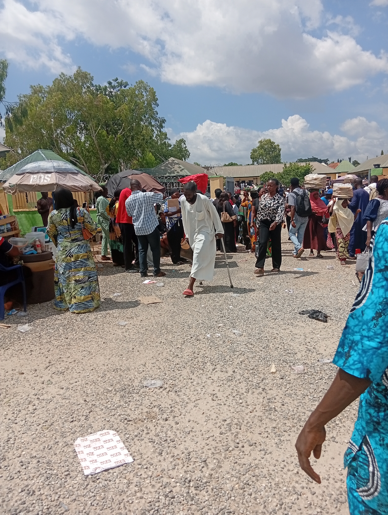 In Jalingo, Nigeria, an outdoor market scene unfolds under a partly cloudy sky. A man, appearing to have a mobility issue, walks with a cane through a gravel-covered area populated by numerous individuals. Several people carry bundles of goods on their heads, suggesting a busy marketplace. Tents and umbrellas provide shade, with various stalls and structures visible in the background. The ground is littered with some debris, including a white paper object with red markings. The overall impression is one of a lively community gathering in a commercial setting.