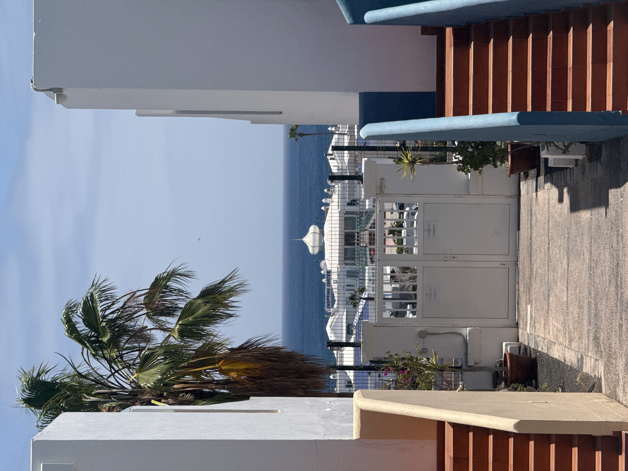 The image captures a scene in Puerto Del Carmen, Spain, featuring a partial view of the ocean and elements of local architecture. The buildings are primarily white with blue and terracotta accents. A palm tree is prominently positioned in the lower left, with its fronds extending towards the center of the frame. In the midground, a gated entrance leads toward a complex of buildings and the ocean beyond. The gates are white, and the complex beyond appears to include a shopping center with clear signage. The ocean stretches to the horizon under a clear, light blue sky. Steps with terracotta-colored risers are visible on the right side of the frame. Shadows suggest the presence of strong sunlight, indicating a bright, clear day.