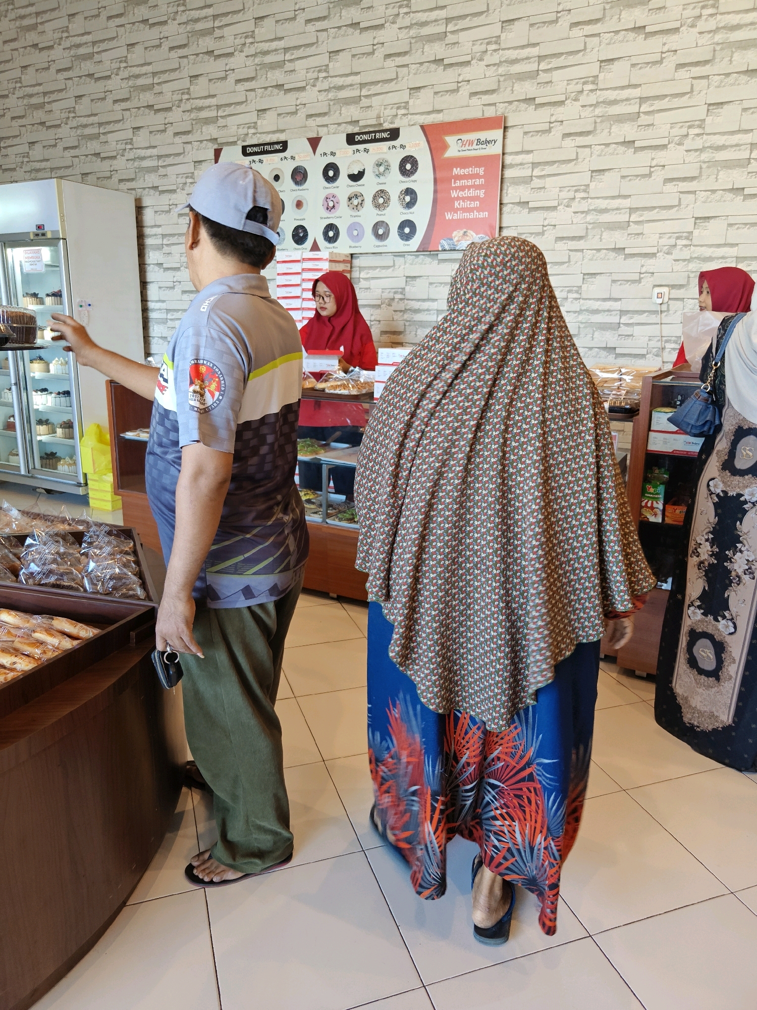 In a bakery in Situbondo, Indonesia, a man wearing a grey baseball cap, a grey and black patterned shirt, and green pants stands near a display refrigerator. He is reaching towards the refrigerator with his left hand. A woman in a red hijab is behind the counter, and another woman is wearing a patterned hijab and a blue and red floral skirt. In the foreground, there are wooden bins filled with bread products. A sign on the wall advertises donuts and bakery services. The floor is tiled in a cream color.