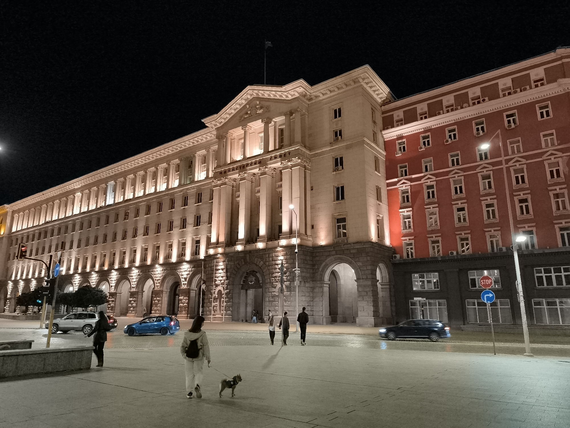 The image depicts a nighttime view of a prominent building in Sofia, Bulgaria, likely the Council of Ministers building. The architecture is classical, with illuminated colonnades and intricate detailing on the facade. To the right, a red-brick building with numerous windows stands adjacent. The scene is a public square, paved with cobblestones.
In the foreground, a woman walks a small dog on a leash. She is wearing light-colored clothing. Behind her, another person is walking away from the camera, also in light-colored attire, with a backpack. Further back, two individuals are walking hand-in-hand, and another person is walking in the same direction.
Several vehicles are visible on the street. A silver SUV is parked on the left, and a blue car is driving away from the viewer. A dark-colored sedan is captured in motion on the right side of the frame, its headlights creating a slight blur.
Traffic lights and streetlights illuminate the area, casting a warm glow on the buildings. A "STOP" sign and a blue directional sign are mounted on a pole. The sky is dark, typical of a nighttime setting. The overall atmosphere is one of urban activity continuing into the evening.
