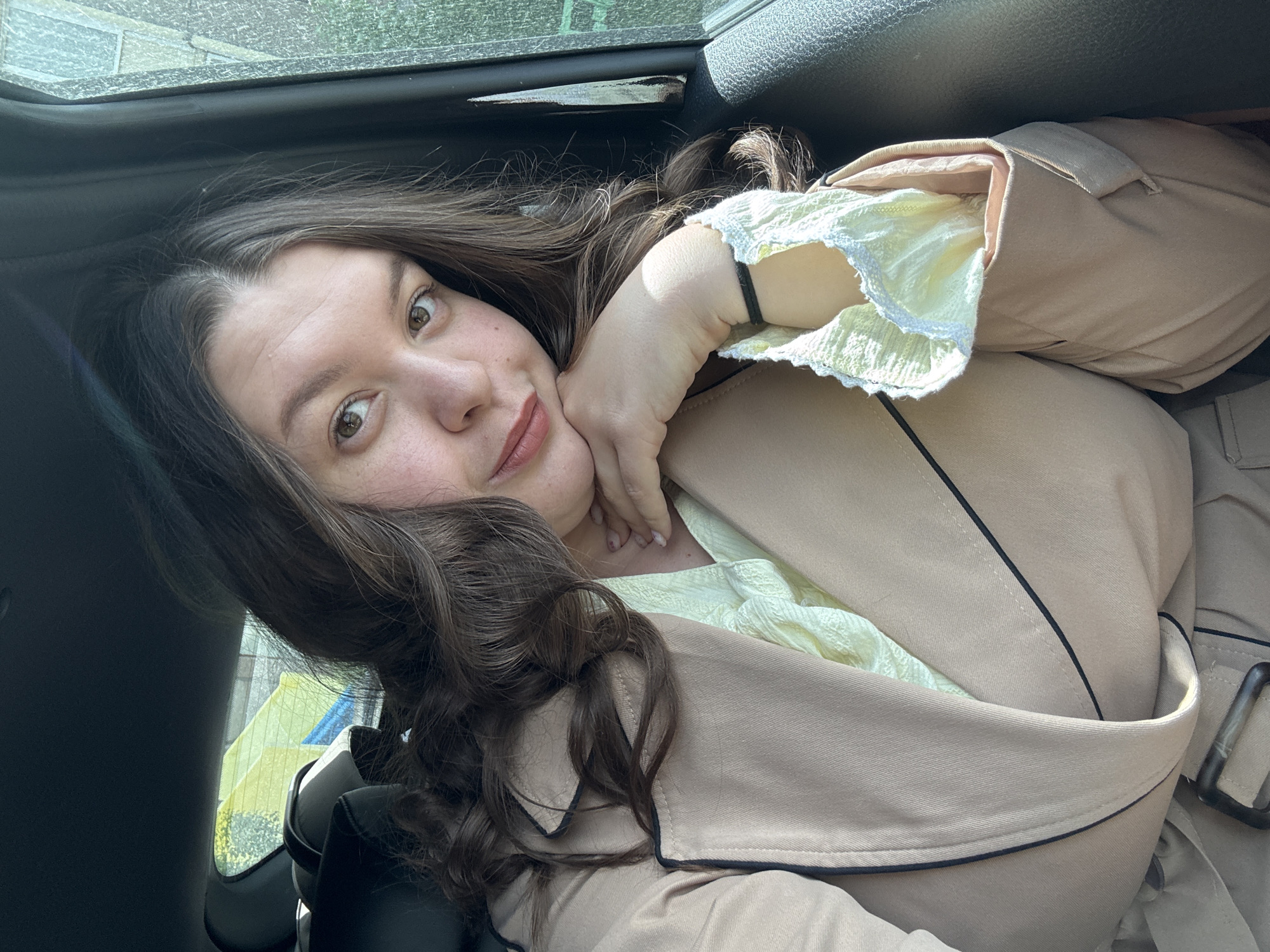 A woman is inside a car. She is wearing a beige trench coat over a light yellow blouse with ruffled sleeves. Her left hand is supporting her chin. The car window is visible above her head. Based on the window reflections, the scene seems to be outside, potentially in a city with buildings and trees. However, definitively identifying the location as Марковский, Russia, is not possible without additional information from the image.