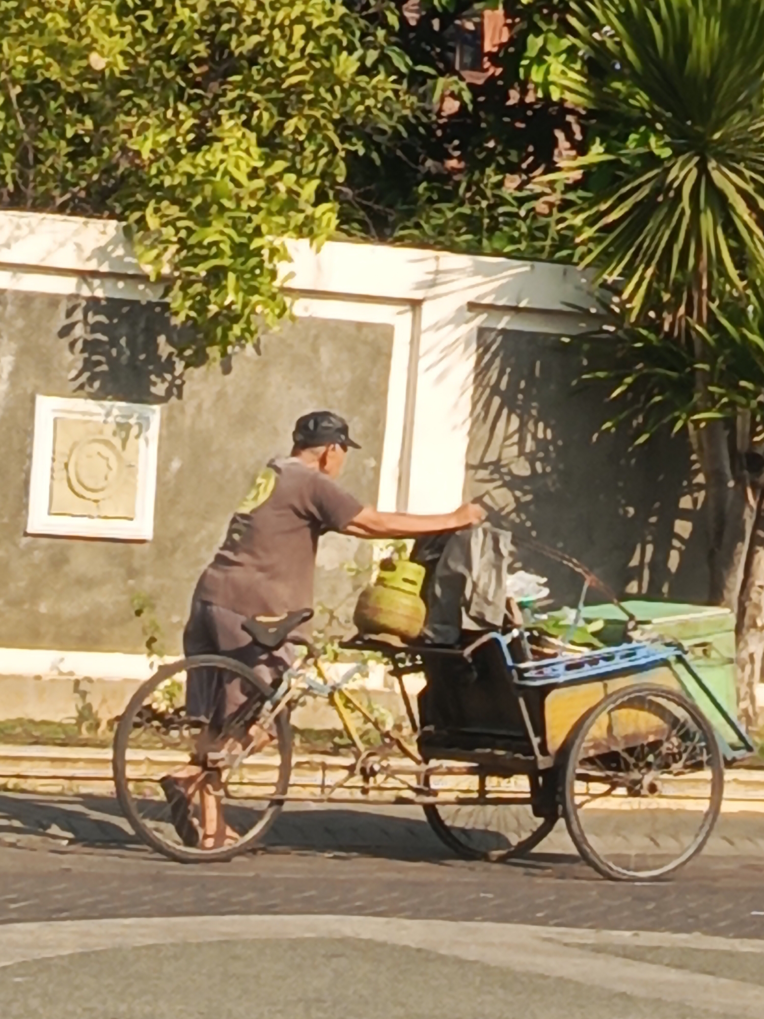 An undated, outdoor, medium-shot photograph captures a man pushing a bicycle with a cart attached to it along a paved road. The man, seen from the side and slightly from behind, wears a dark t-shirt, dark shorts, and a black baseball cap. He is pushing the cart with his right hand. The cart, which appears to be a modified bicycle with two wheels at the rear and a third in the front to support the cargo area, carries a green gas cylinder, some green produce, and other items covered with a dark tarp. The bicycle has a single rear wheel and a front wheel that is not fully visible. The cart has a yellow frame and a green cargo box. The background consists of a textured wall with a decorative square element and foliage, including a palm tree on the right. The lighting suggests it is daytime, with shadows cast on the wall. The scene is presumed to be in Boro, Indonesia, given the context.