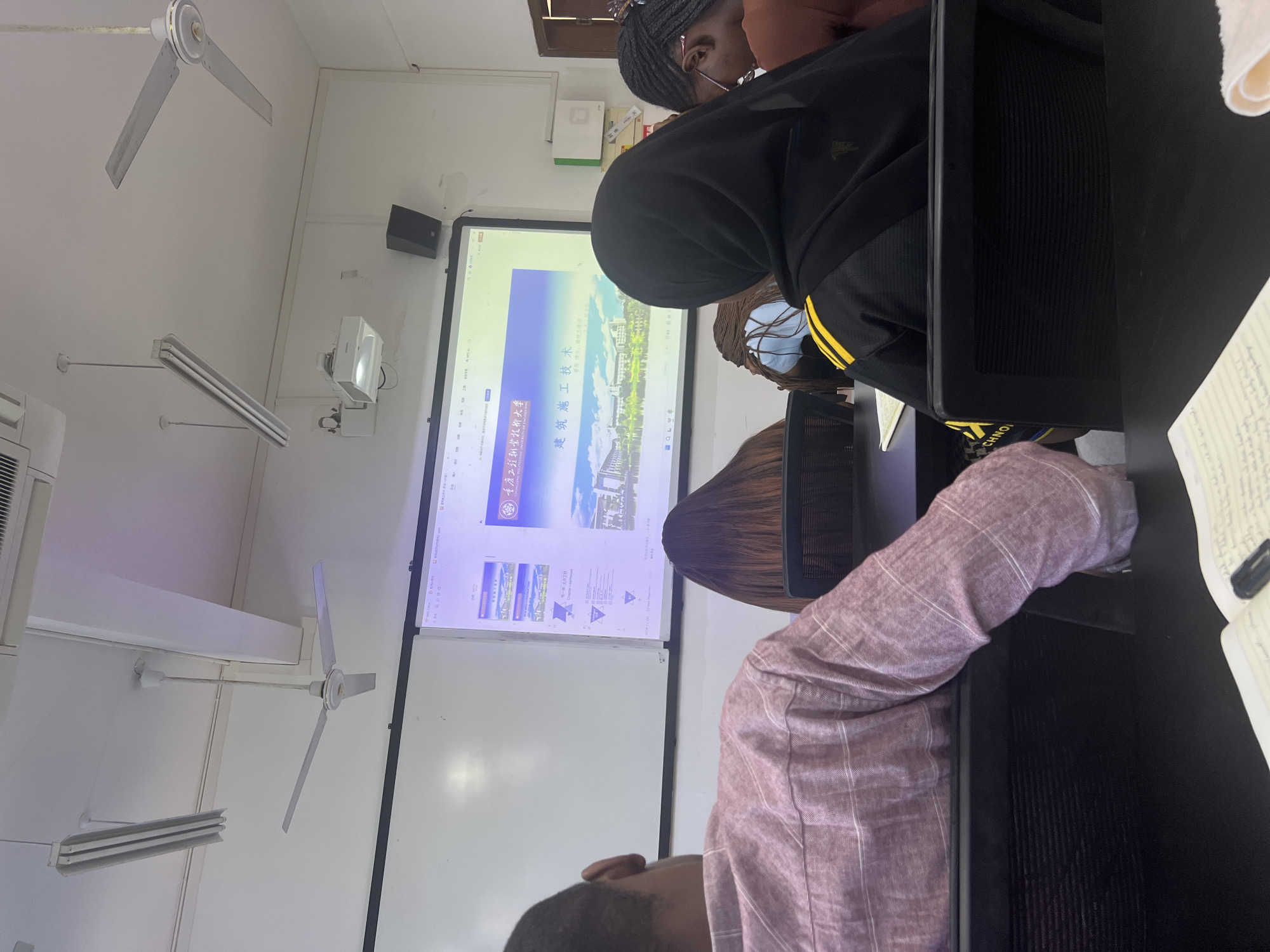 This is a medium shot of a classroom in Jiangjin, China, where students are attending a lecture. The projector screen displays a presentation with Chinese text and images of university buildings. In the foreground, three students are visible from behind. The student on the left is wearing a pink and white striped shirt, the student in the center is wearing a black hijab and a black jacket with yellow stripes, and the student on the right is wearing a brown shirt and has braided hair. The classroom environment appears functional, with ceiling fans and fluorescent lighting. The overall scene suggests an academic setting with an active lecture in progress.