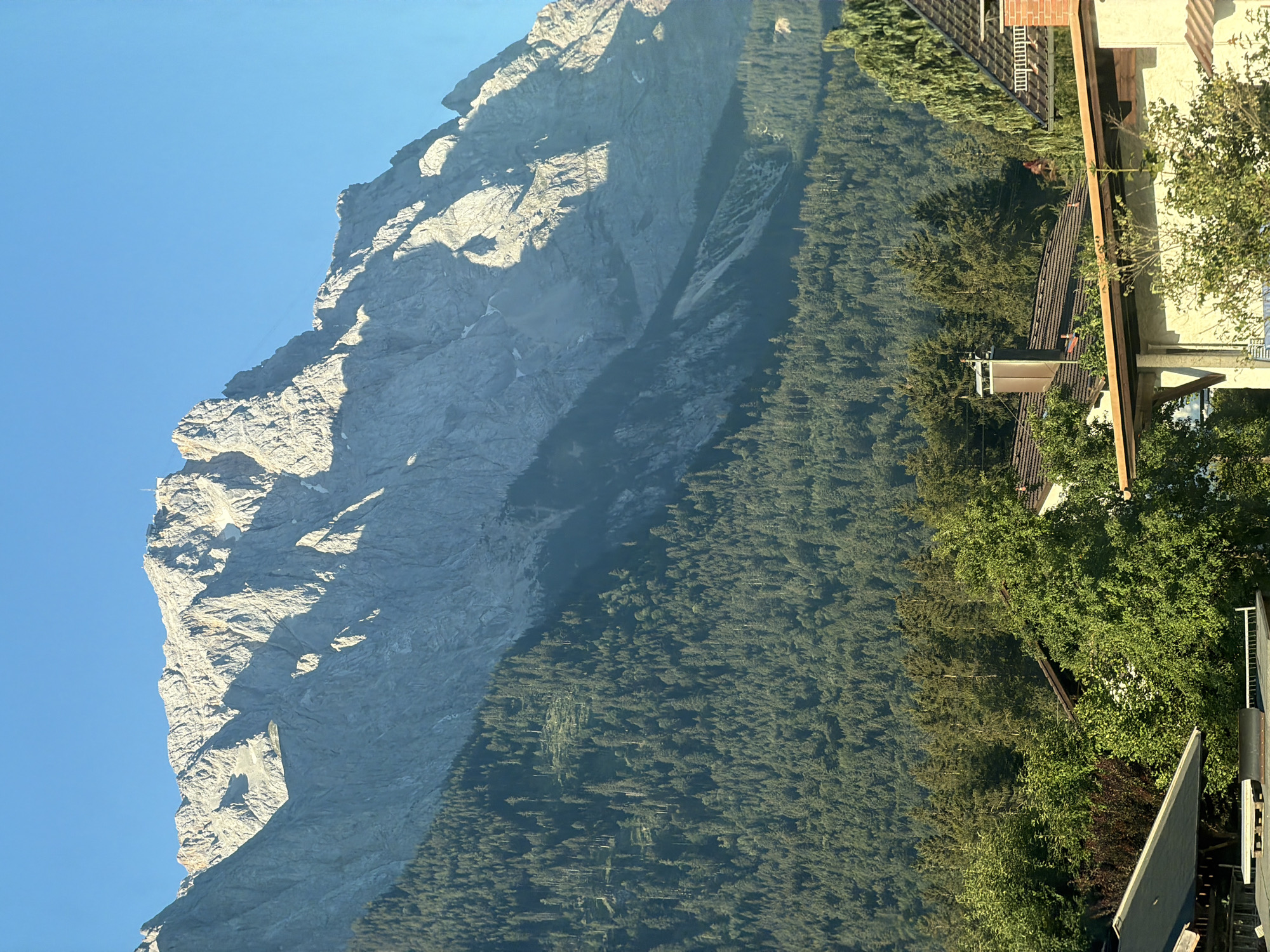 The image depicts a scenic view of the Zugspitze mountain range in Grainau, Germany. The upper portion of the frame is dominated by the imposing, rocky peaks of the Zugspitze, bathed in the warm, golden light of the sun, casting deep shadows across its rugged face. A building or structure is visible at the summit, and a cable car line extends diagonally across the mountain's slope. Below the mountain, a dense, verdant forest covers the lower slopes, with a clear blue sky overhead. In the foreground, the rooftops of houses and trees are visible, suggesting the presence of a village or settlement nestled at the foot of the mountains. The scene is a daytime exterior shot, showcasing the natural beauty of the Bavarian Alps.