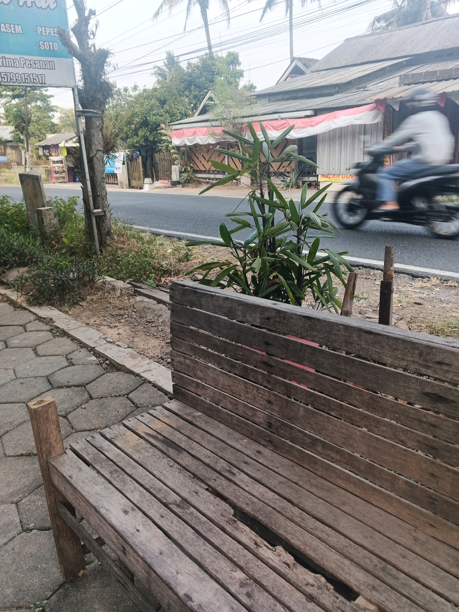 The image depicts a street scene in Ungaran, Indonesia, characterized by a weathered wooden bench in the foreground, a paved walkway, and a road. A person on a motorcycle is blurred in motion on the road, suggesting a sense of movement. The background features buildings with corrugated roofs, trees, and vegetation. A sign is visible on the left, displaying text in Indonesian, with the partial text "ASEM PEPE" and "TERIMA PESANAN" being legible. The overall impression is that of a typical street view in a Southeast Asian town.