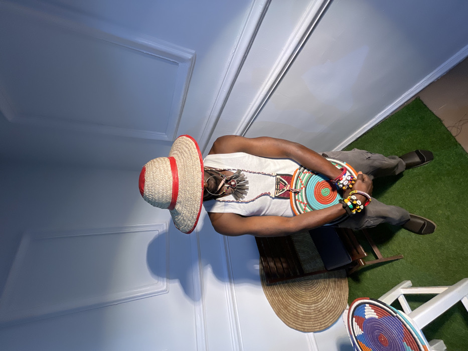 An individual stands centrally in an indoor setting in Jos, Nigeria. The subject wears a white sleeveless top, dark grey cargo pants, and dark closed-toe shoes. A wide-brimmed straw hat with a red band and red brim trim obscures the upper face. Multiple necklaces are visible around the neck, including one with dark, tubular elements and another with a red and dark patterned triangular pendant. Both wrists display several multi-colored beaded bracelets, some featuring cowrie shells. The individual holds a circular, shallow woven platter with concentric bands of orange, light blue, teal, green, white, and dark colors against their torso. The environment features light blue walls with white decorative panel molding and a green artificial turf floor. To the left of the individual, a dark wooden chair with a dark seat cushion is present. Behind the chair, a large, round, flat, beige woven mat leans against the wall. A partial view of a white stool or small table in the lower-left foreground supports a smaller, colorful woven basket. Strong directional lighting from the left casts a defined shadow of the figure and hat onto the wall.