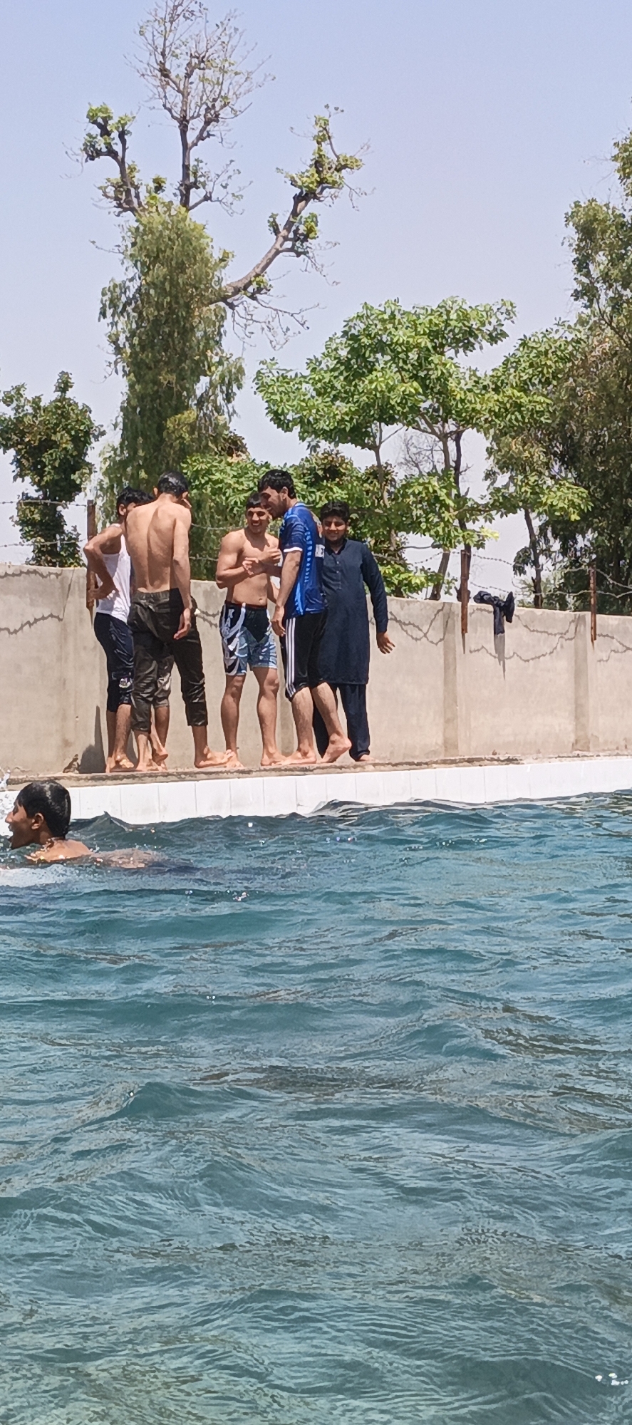 A group of five young men stand on the edge of a swimming pool. One man swims in the pool, his head and shoulders visible above the water. The men on the edge of the pool are dressed in various stages of swimming attire. The background includes a concrete wall topped with barbed wire and greenery with trees. The setting appears to be an outdoor swimming pool area. This is a candid shot that can be associated with the city of Samundri, Pakistan.