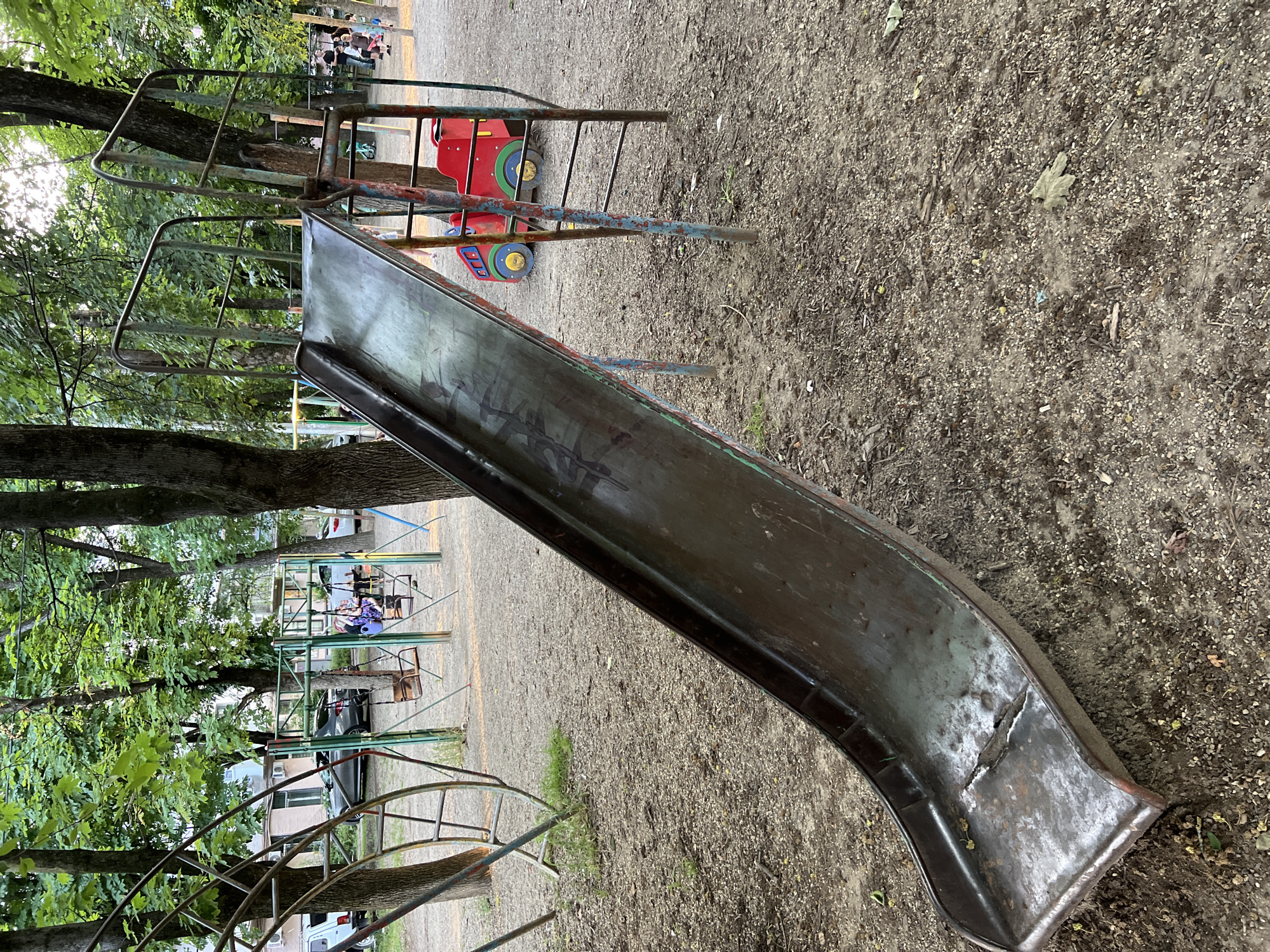 This is an outdoor image of a children's playground in Krasnodar, Russia. In the foreground is a metal slide, showing signs of wear and tear, including rust and a hole near the bottom. The slide is supported by metal poles. To the left of the slide, part of a metal climbing structure is visible. In the background, there are trees with green leaves, suggesting it is springtime or summer. There are also swings and other playground equipment. Several cars are parked in the background, and some buildings can be seen through the trees. A few people are present in the background, but their activities are not clearly discernible. The ground is covered with gravel and dirt, with some scattered leaves and debris. A red toy vehicle with yellow wheels is also visible near the slide.