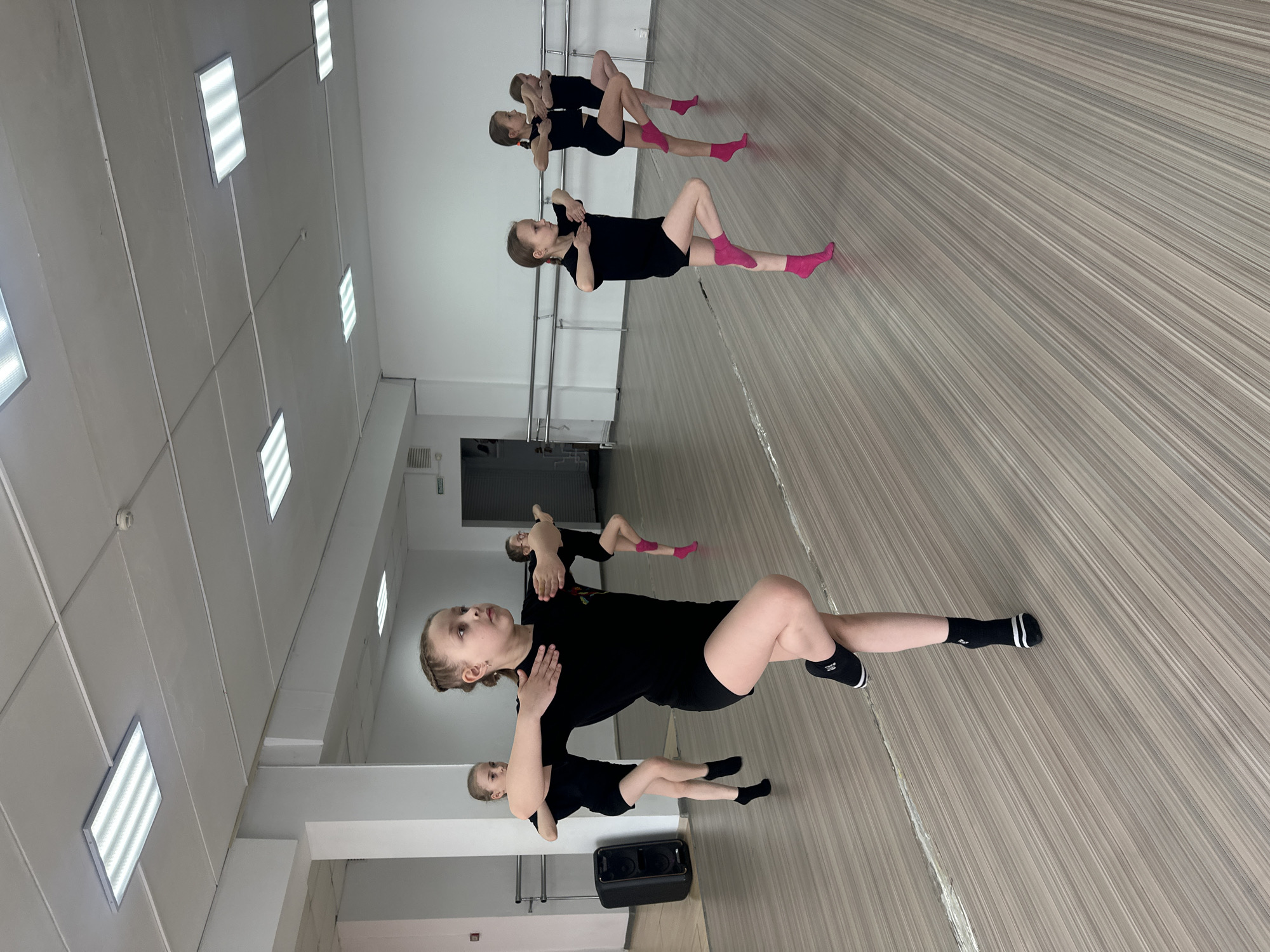 This media file captures a group of young girls performing a floor exercise in what appears to be a dance studio. The image is rotated 90 degrees clockwise, making the subjects appear to be on a wall, but they are in fact lying on their backs on a floor.
The subjects are approximately five young girls, likely pre-teens, dressed in uniform black short-sleeved tops and black shorts. Their footwear varies, with some wearing bright pink ankle socks and others wearing black ankle socks with white stripes. They are positioned in a diagonal line across the room, lying supine with their knees bent and arms bent with hands near their chest or shoulders, indicating a specific dance or exercise routine. Their facial expressions convey focus and concentration.
The setting is a bright, indoor dance studio. The floor is light-colored wood or laminate with a distinct striped pattern. One wall is covered with large mirrors, reflecting the studio space and the girls. A ballet barre runs along the mirrored wall. The ceiling features multiple rectangular fluorescent light panels, providing strong, even illumination throughout the room. In the foreground, a black portable speaker is visible on the floor. In the background, a dark door or shutter is partially seen. There is no clearly legible text visible within the frame. The scene suggests a structured practice or training session in Votkinsk, Russia.