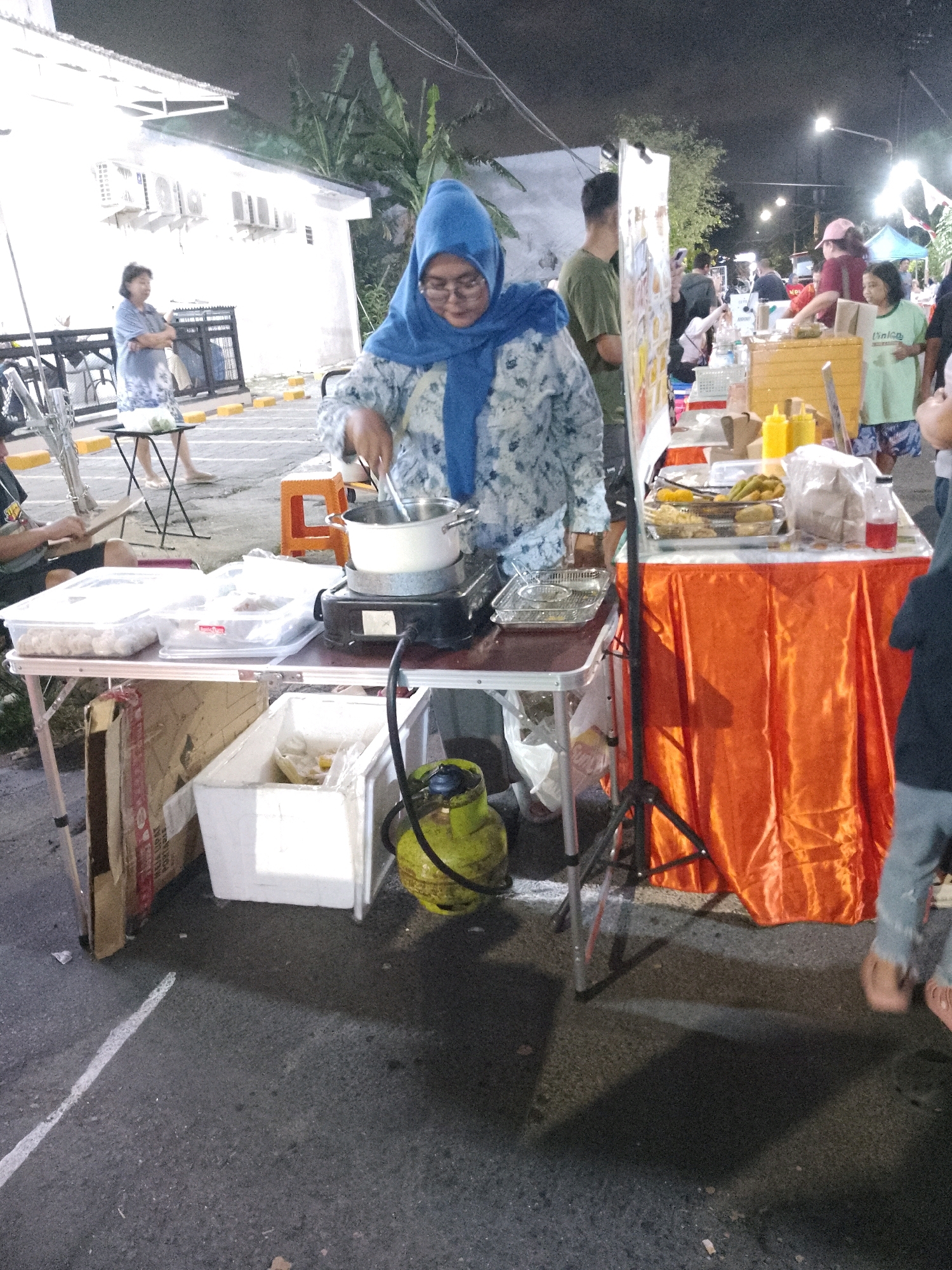 The image depicts an outdoor nighttime scene at a street food stall in Surabaya, Indonesia. A woman wearing a blue hijab and patterned blue and white clothing is preparing food, stirring a pot on a portable stove. In front of her is a table laden with food items, containers, and condiments, draped with an orange tablecloth. To her left, a gas cylinder is visible, as is a white cooler box. In the background, other people are visible at various food stalls and walking around. Buildings and trees are also present in the dimly lit environment, illuminated by streetlights. The ground appears to be asphalt, typical of a street setting.