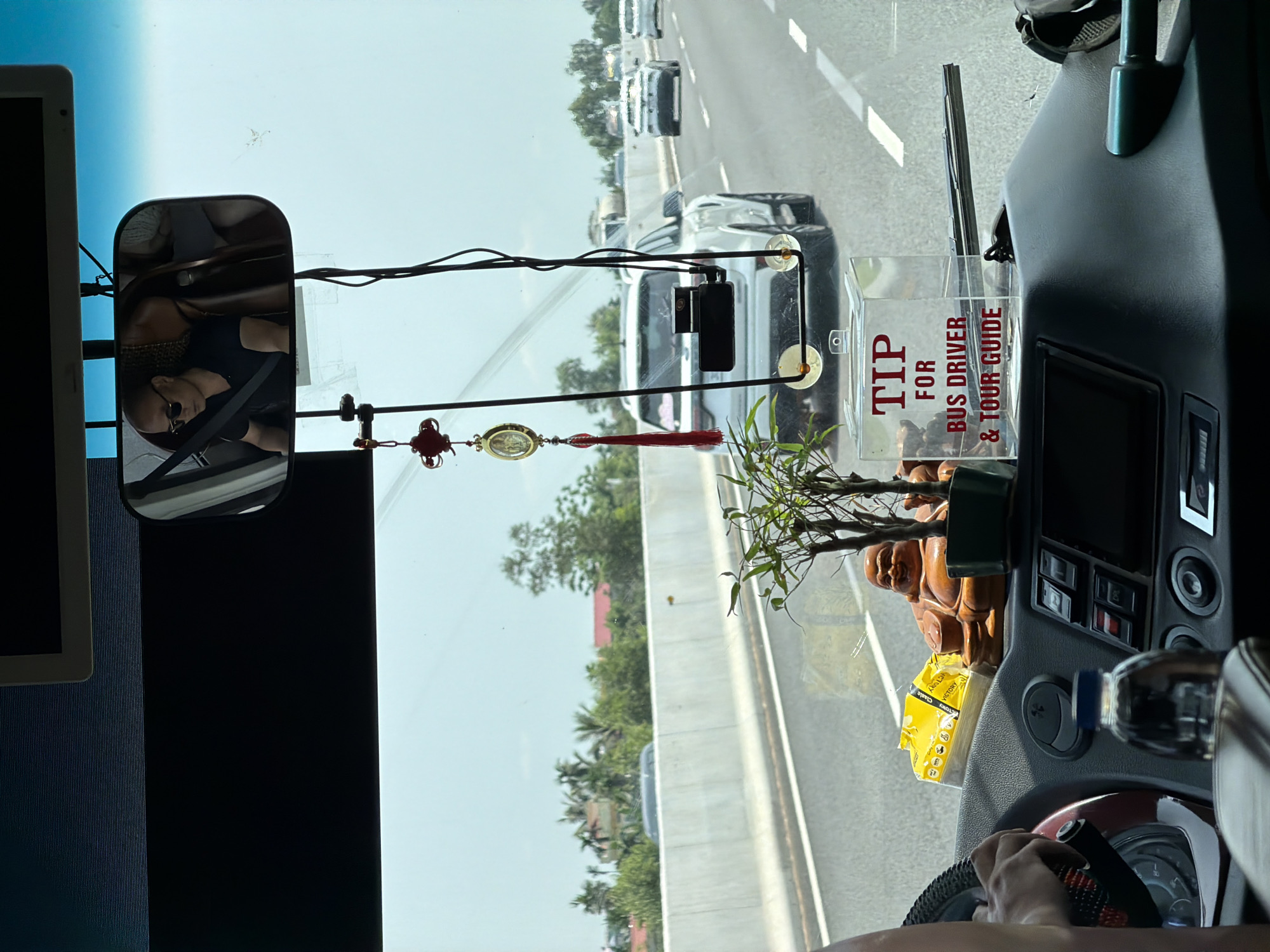The image captures an interior view from a vehicle, likely a bus or tour van, looking out onto a multi-lane highway in Hải Phòng, Vietnam, on a bright, clear day.
In the immediate foreground, a driver's hands are visible on the steering wheel at the bottom right. The dashboard features various controls, a small display, and is adorned with several items: a water bottle, a yellow package (possibly "VICTORY" brand), a brown Laughing Buddha statue, and a small potted plant. A prominent clear acrylic box is placed on the dashboard, with red text stating "TIP FOR BUS DRIVER & TOUR GUIDE." Wires run across the upper portion of the windshield, from which a decorative red and gold tassel hangs.
Looking out the windshield, a rearview mirror on the left reflects a person wearing sunglasses. A dash camera is also visible, mounted high on the windshield. The highway ahead shows other vehicles, including cars and trucks, moving along. A concrete barrier separates the lanes. Beyond the road, green trees, including palm trees, and some distant structures or houses can be seen under a bright, blue-white sky, indicating sunny weather. The overall scene conveys a journey in progress during daytime.