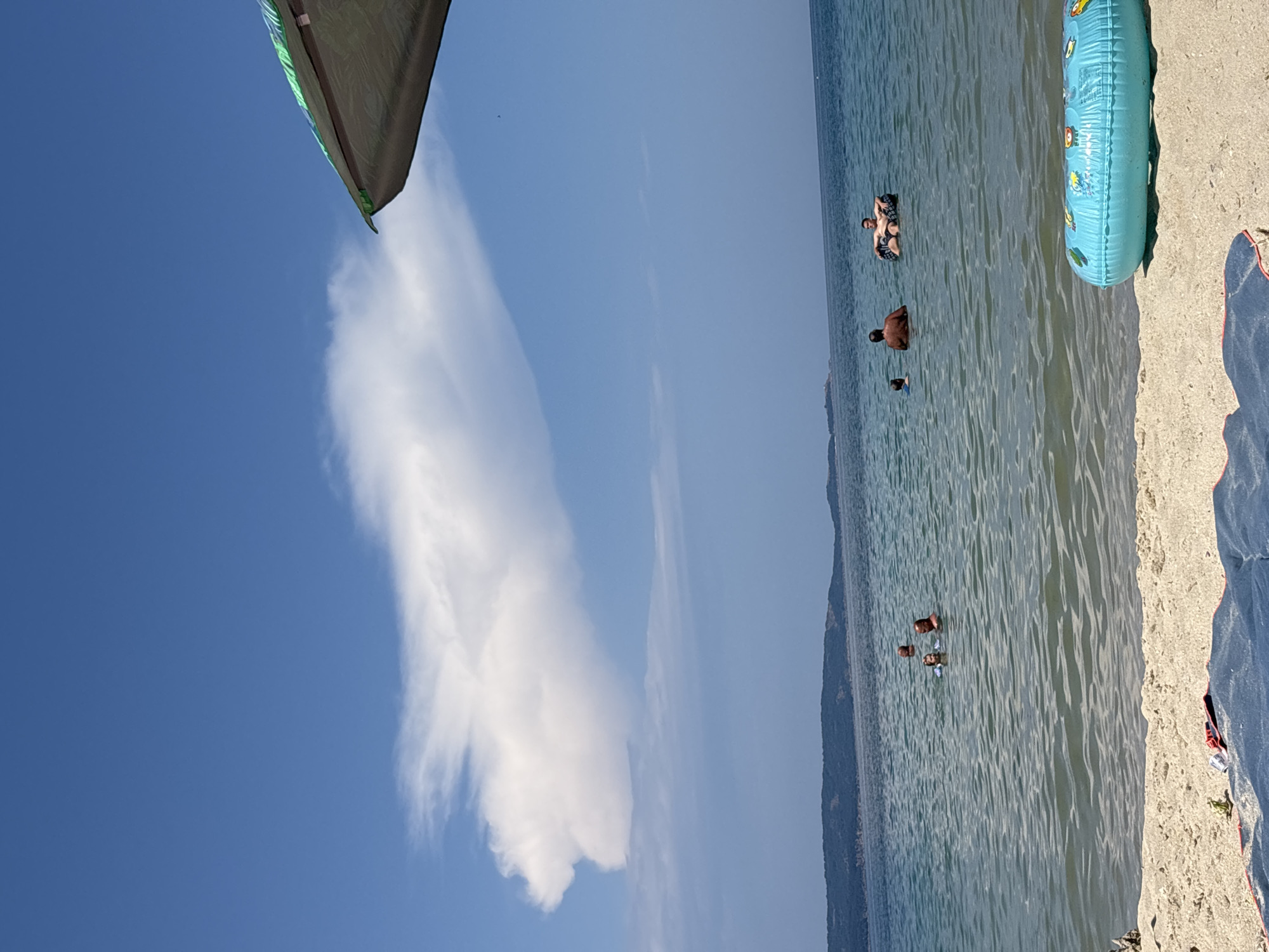 This is a daytime, eye-level, wide shot of a beach and the sea in Kiten, Bulgaria. The sky is a clear blue with a large, puffy white cloud. The sea is calm with gentle waves. In the water, several people are visible: a few are swimming, one is floating on an inflatable tube, and another is partially submerged. On the sandy shore, a blue inflatable ring with cartoon animals is lying on the sand, along with a blue towel and a small green object. In the background, a low, tree-covered hill is visible across the water. A portion of a beach umbrella is visible in the upper right corner of the frame.