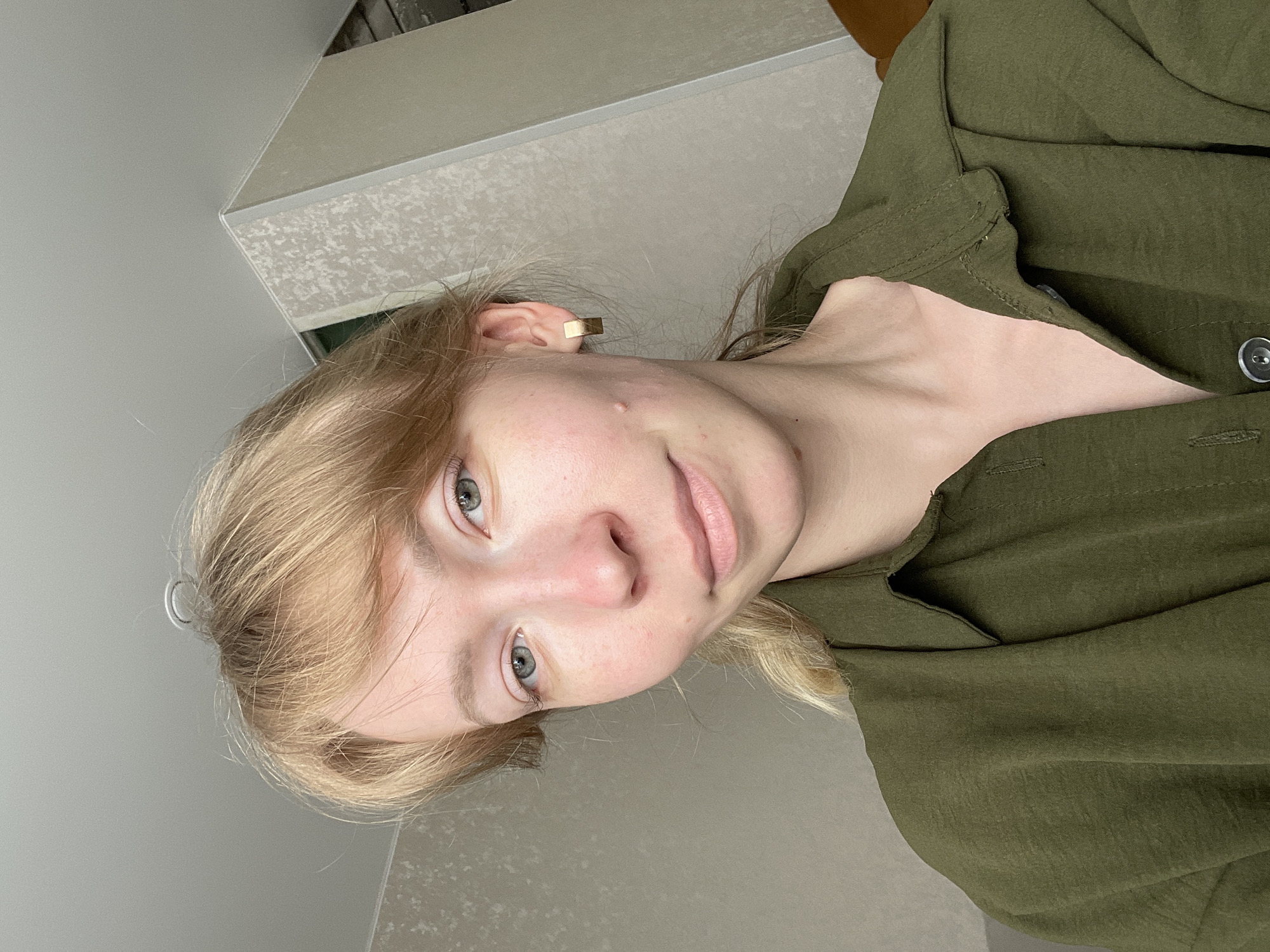 A medium, vertical close-up shot shows a fair-skinned person with blonde hair, light blue eyes, and a pink lip color. The person is wearing a green, button-down shirt. A gold, rectangular earring is visible in the left ear. The background consists of a gray wall and a textured beige surface, potentially a counter or table. The scene is likely indoors. There is no explicit indication of the location being Perm, Russia in the image.