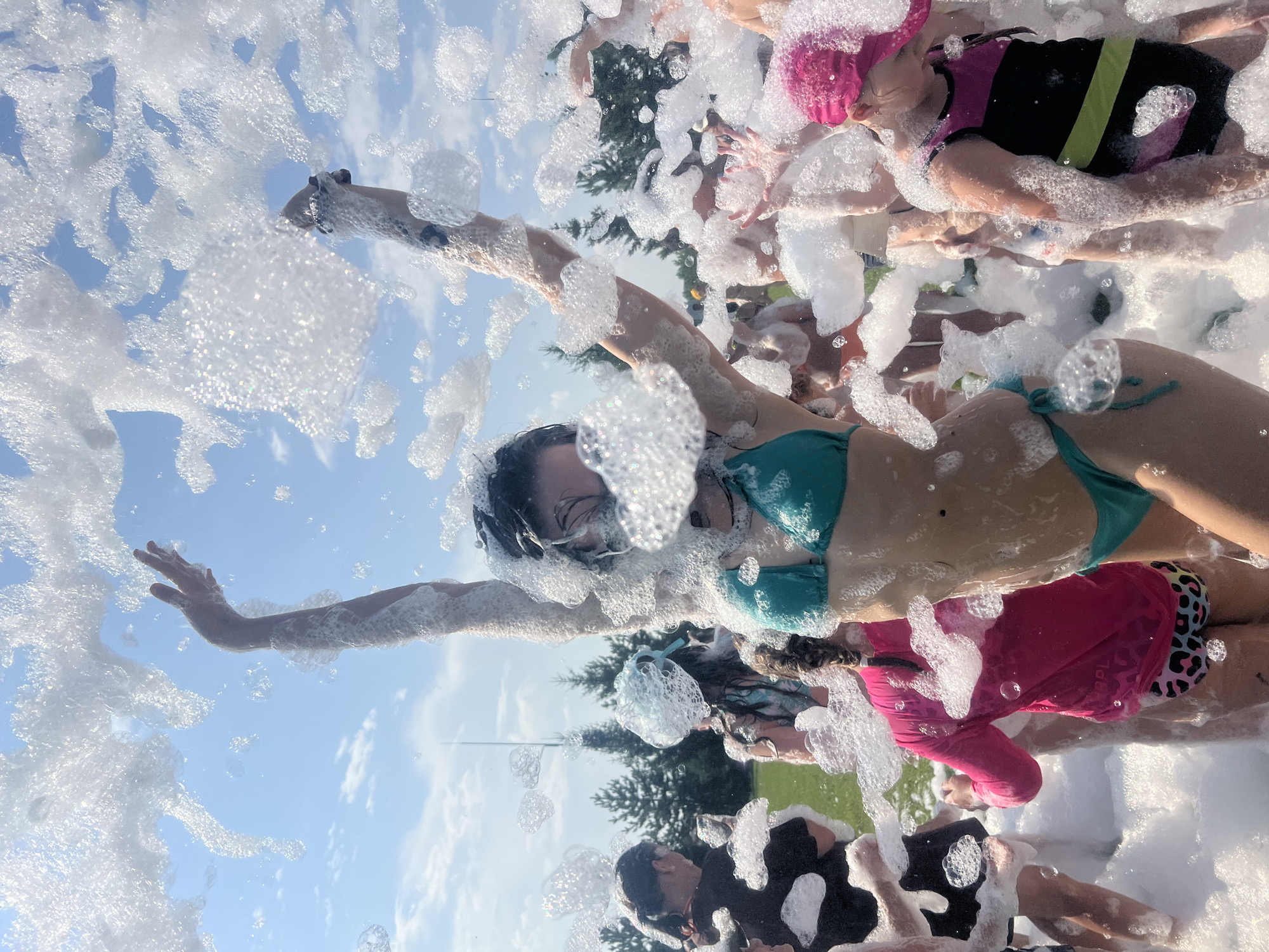 In a sunlit outdoor setting, likely at Емельяновское озеро, Russia, a group of people, predominantly young women and girls, are immersed in a foam party. The foreground is dominated by a woman in a turquoise bikini, her arms raised joyfully amidst a cascade of white foam. Her face is partially obscured by the foam, but her energetic pose suggests exhilaration. Behind her, other individuals are visible, some also in swimwear, interacting with the foam. In the background, trees and a bright blue sky with scattered clouds suggest a warm, pleasant day. The overall atmosphere is one of carefree fun and celebration, with the foam creating a playful and dynamic visual effect.