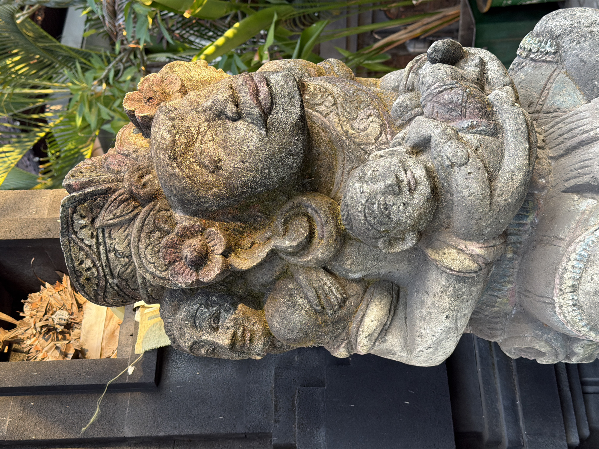In Mengwi, Indonesia, a weathered stone sculpture depicts a group of figures in a close embrace. The sculpture is composed of at least four distinct faces, each carved with individual features and expressions. The primary figure, a large central face, gazes slightly upwards with closed eyes and pursed lips, adorned with what appears to be a crown or headdress decorated with floral motifs. To the left and slightly behind this central figure, another face with open eyes and a gentle smile is visible, cradling a smaller, round object in its arms. Below this, a third, smaller face with closed eyes and a subtle smile peeks out from the embrace, also holding a rounded object. A fourth, more abstract face is partially visible at the very top of the sculpture. The stone itself shows signs of age, with a rough texture and patches of discoloration. The sculpture is positioned outdoors, with a dark, textured wall serving as a backdrop to the left. To the right, lush green foliage, including palm fronds, is visible, illuminated by warm, directional light, suggesting it is either early morning or late afternoon. The overall scene evokes a sense of tradition and artistic heritage.