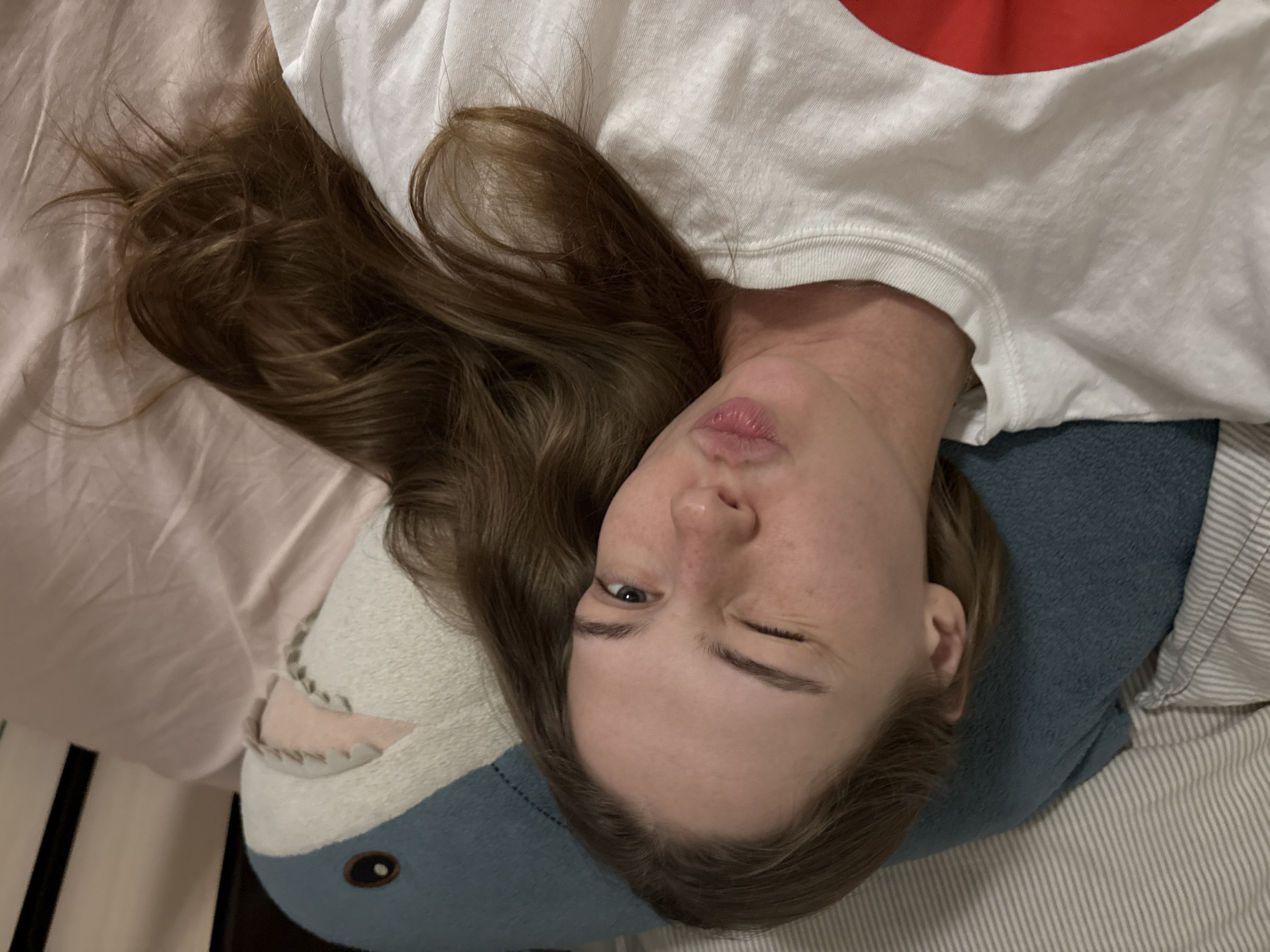 The image is a close-up, top-down selfie of a young woman lying down, possibly in a bed. She is winking and puckering her lips, suggesting a playful or flirtatious pose. Her long, light brown hair is spread across her shoulders and the surrounding pink fabric, which appears to be bedding. She is wearing a white t-shirt with a large red circular design on the left side of the chest. Behind her is a large plush toy resembling a blue shark. The background includes striped bedding and the pink fabric. There is no indication of the city of Perm, Russia, or any other specific location.